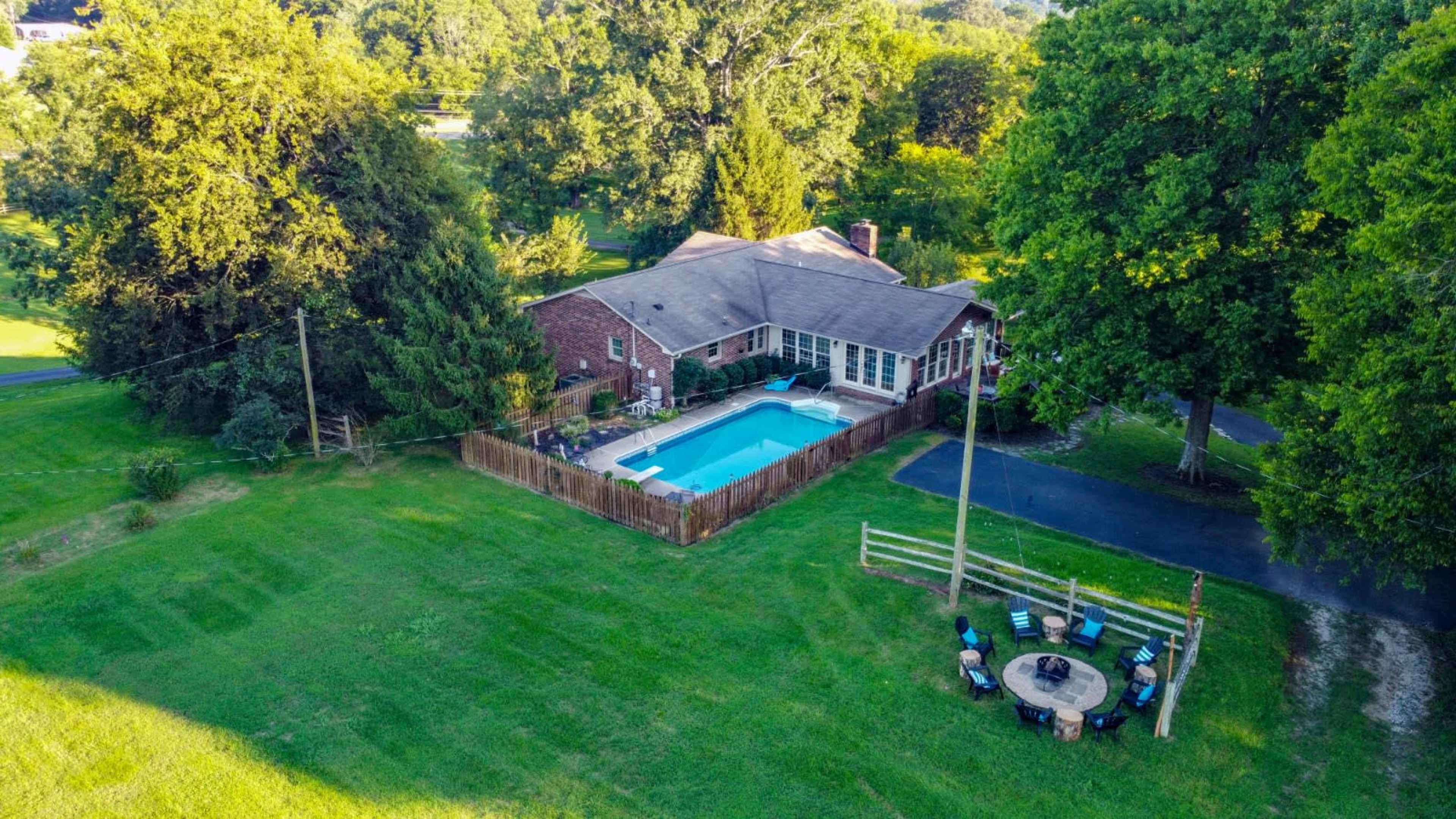 The image shows a single-story brick house with a swimming pool in the backyard, surrounded by a wooden fence and dotted with trees.