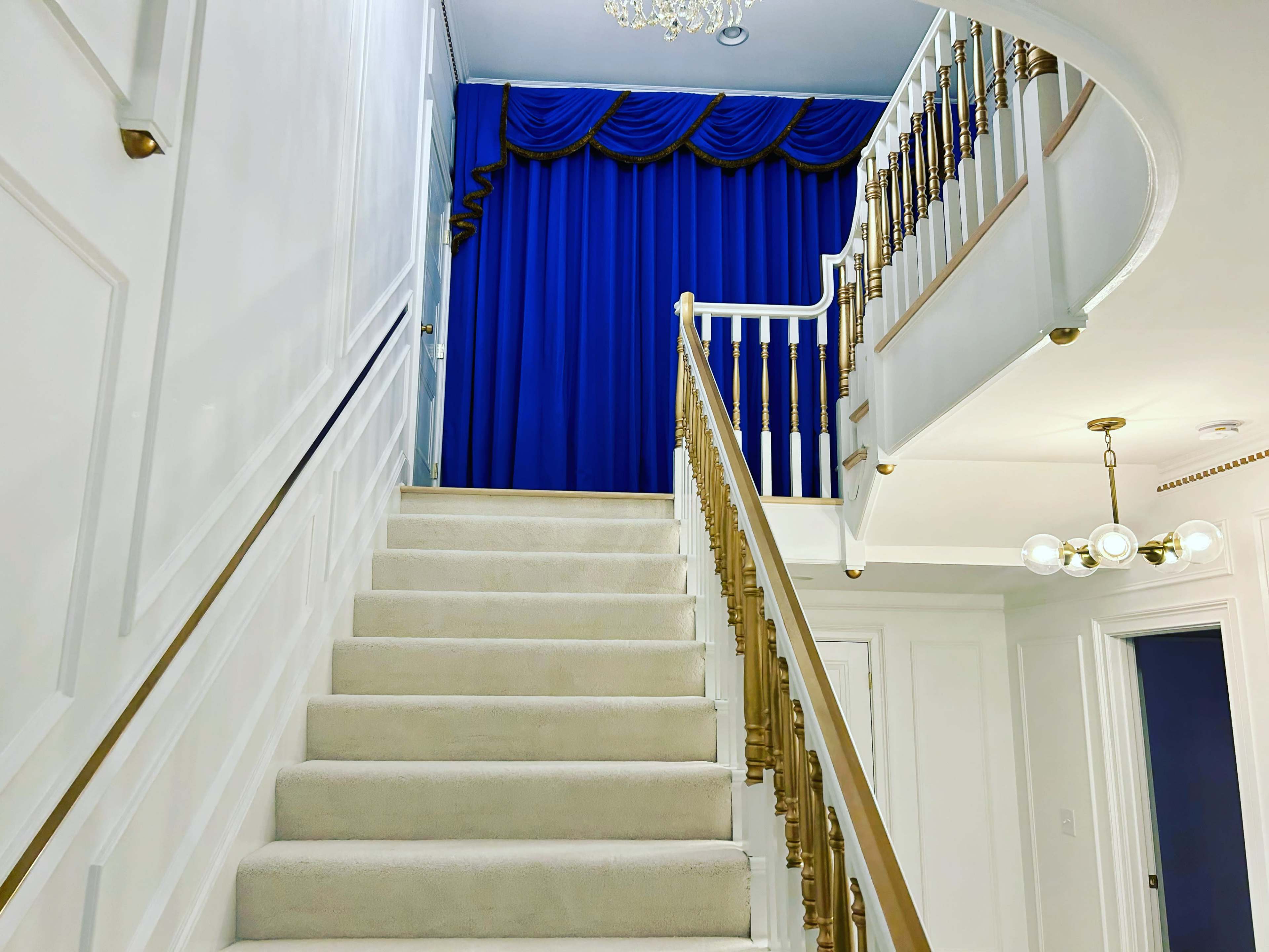 The image shows a staircase leading to a landing with a blue curtain and a chandelier above.
