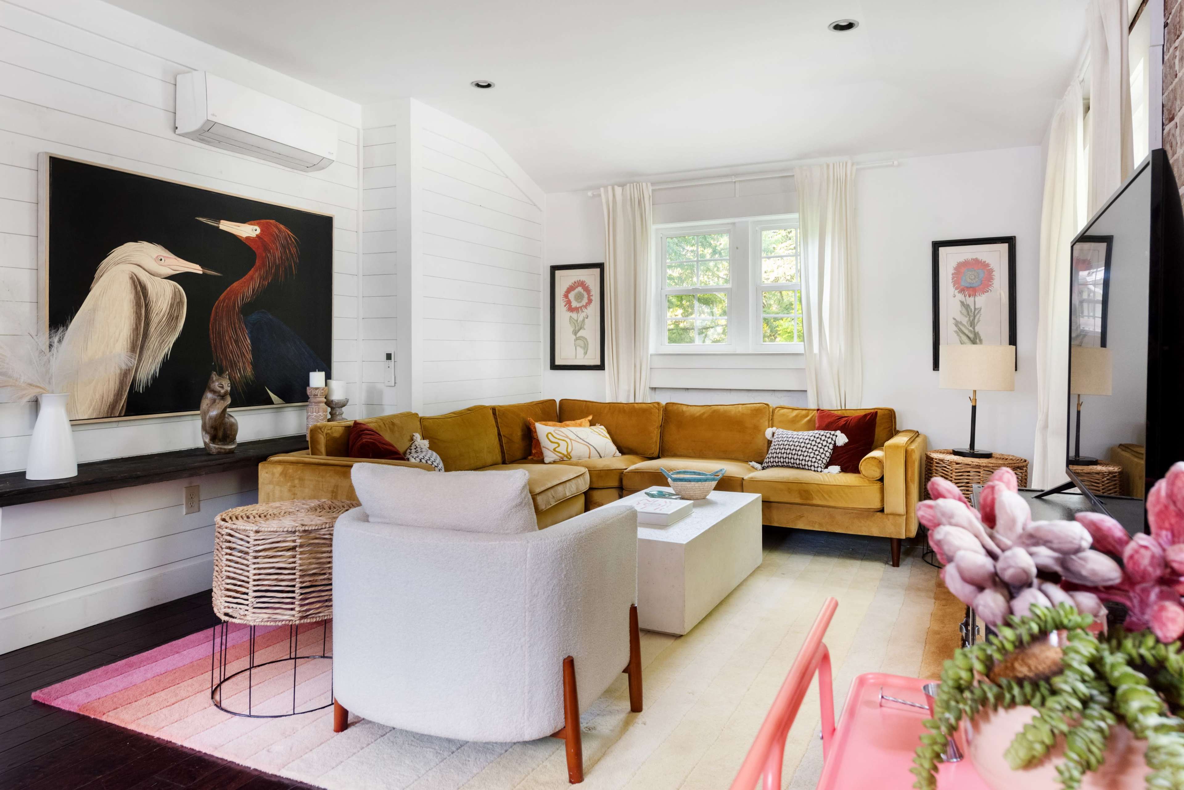 The living room features a yellow sectional sofa, a white armchair, and a large artwork of two birds on the wall, with decorative plants and light-colored curtains.