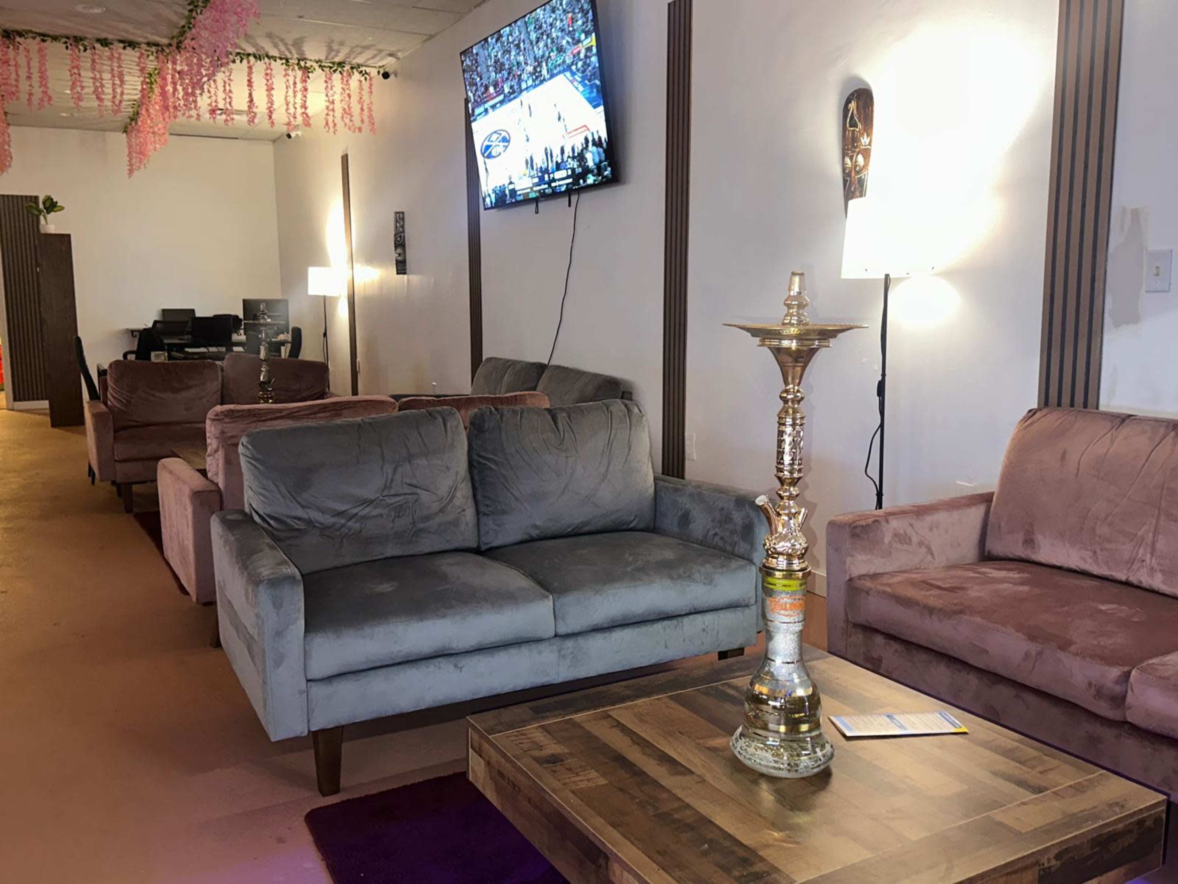 The image shows a lounge area with two couches, a wooden table with a hookah, and a television mounted on the wall displaying a sports game.