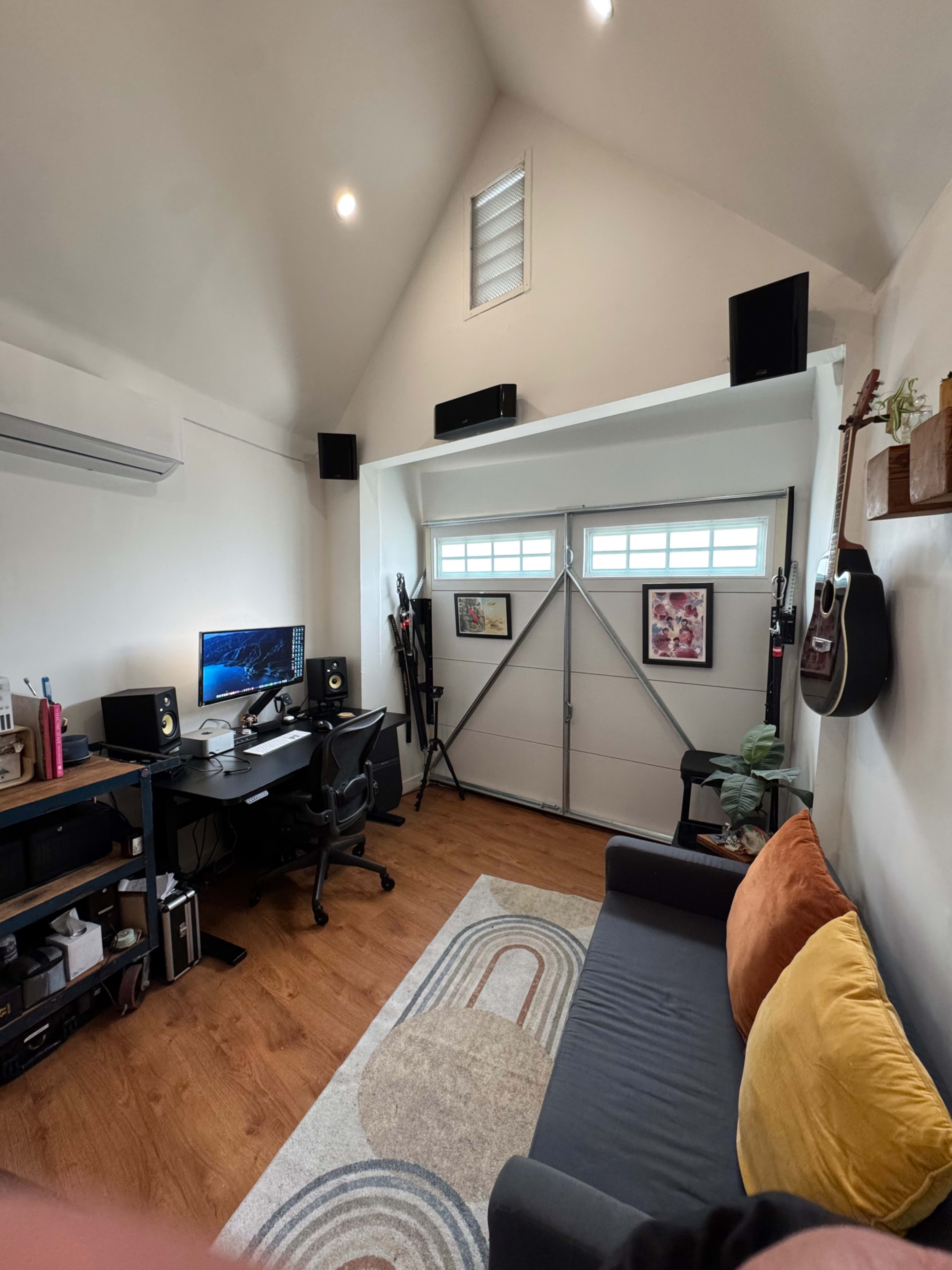 The image shows a small home office featuring a dark desk with a computer setup, a black chair, and a couch with yellow and orange cushions, situated near a garage door with windows.