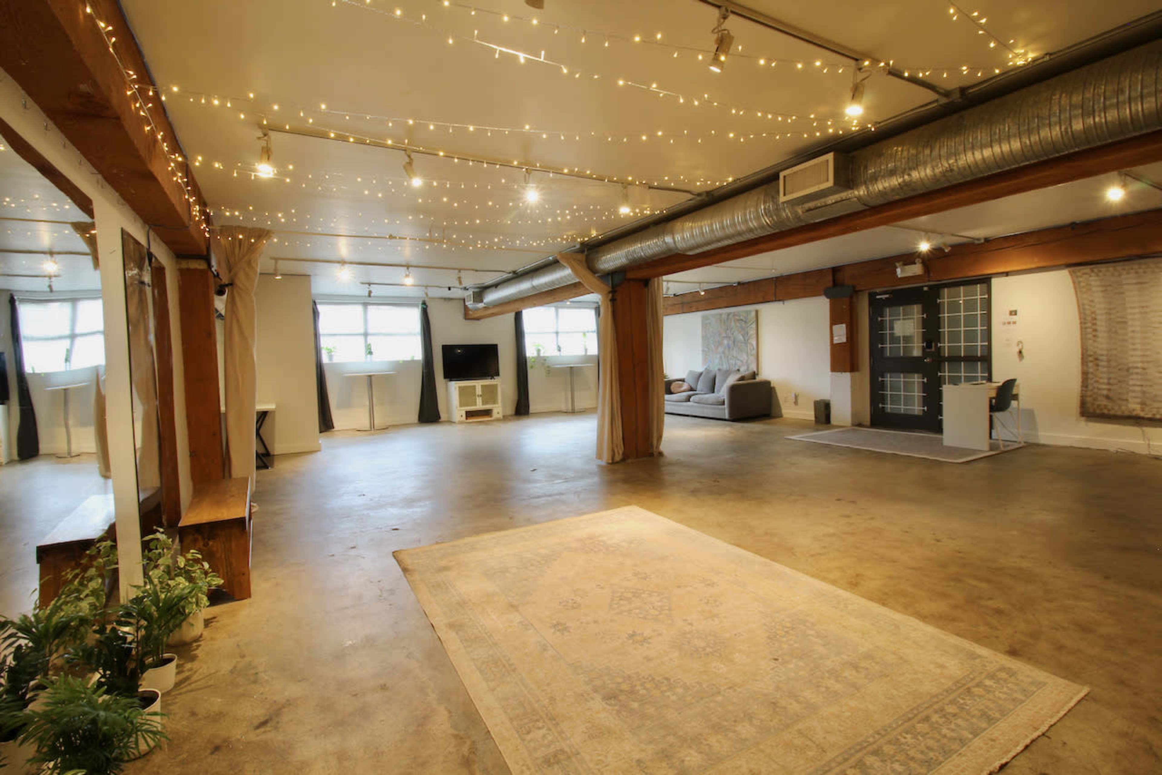 The image shows a spacious, well-lit interior with a concrete floor, string lights across the ceiling, and a few pieces of furniture including a couch and a television.