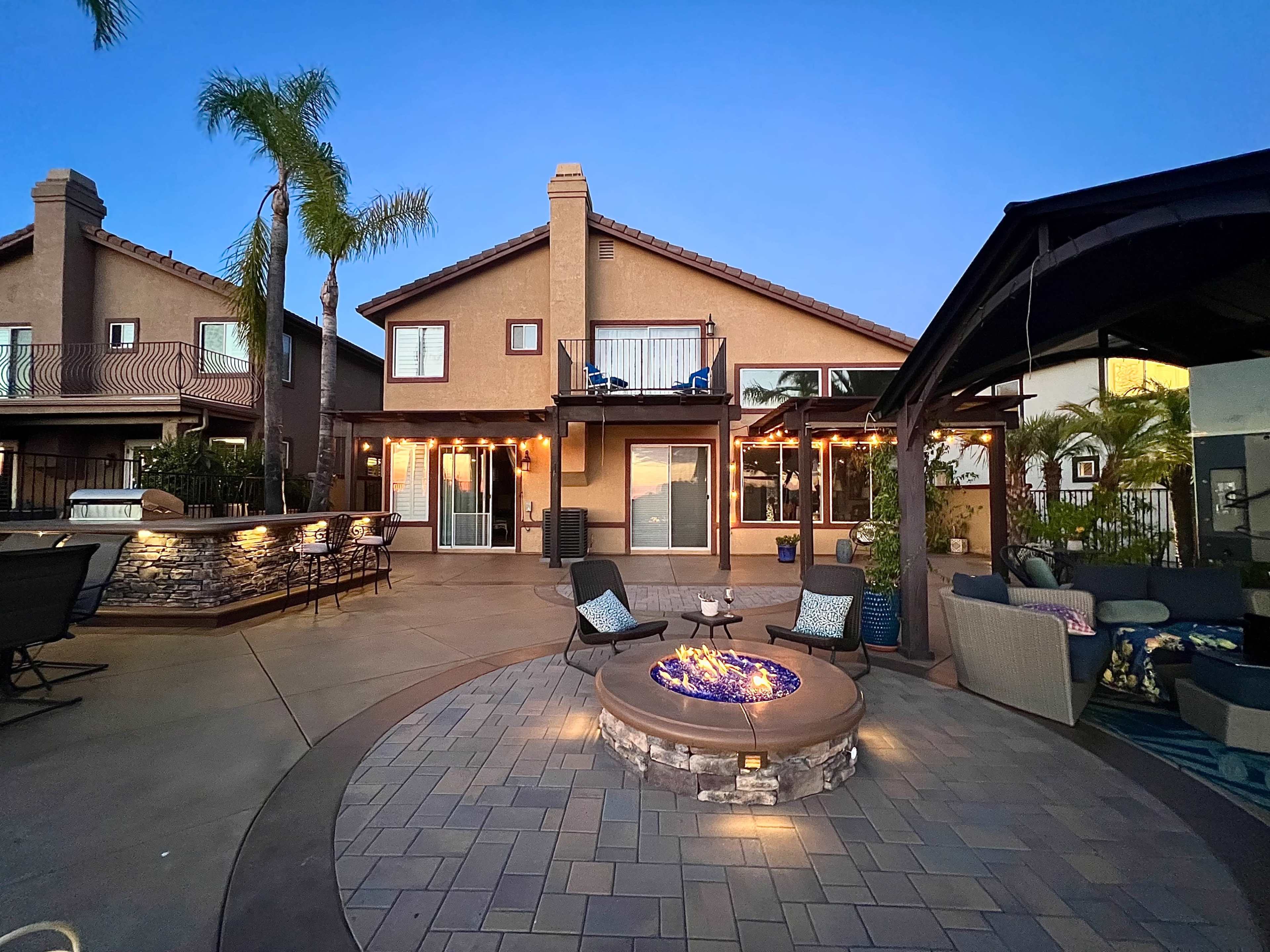 A two-story house with outdoor seating and a fire pit is illuminated in the evening light, surrounded by palm trees and a patio area.