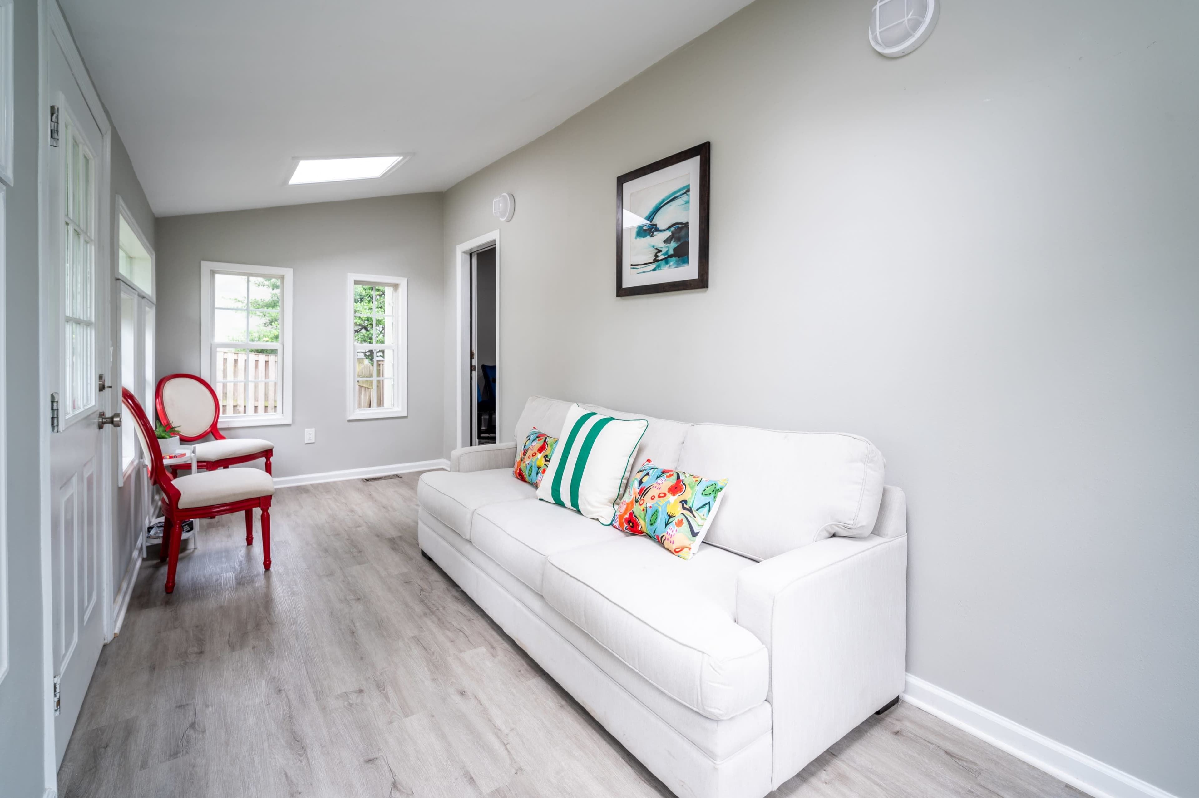 A light-colored room with a white sofa and a red chair near two windows, featuring a white wall and wooden flooring.