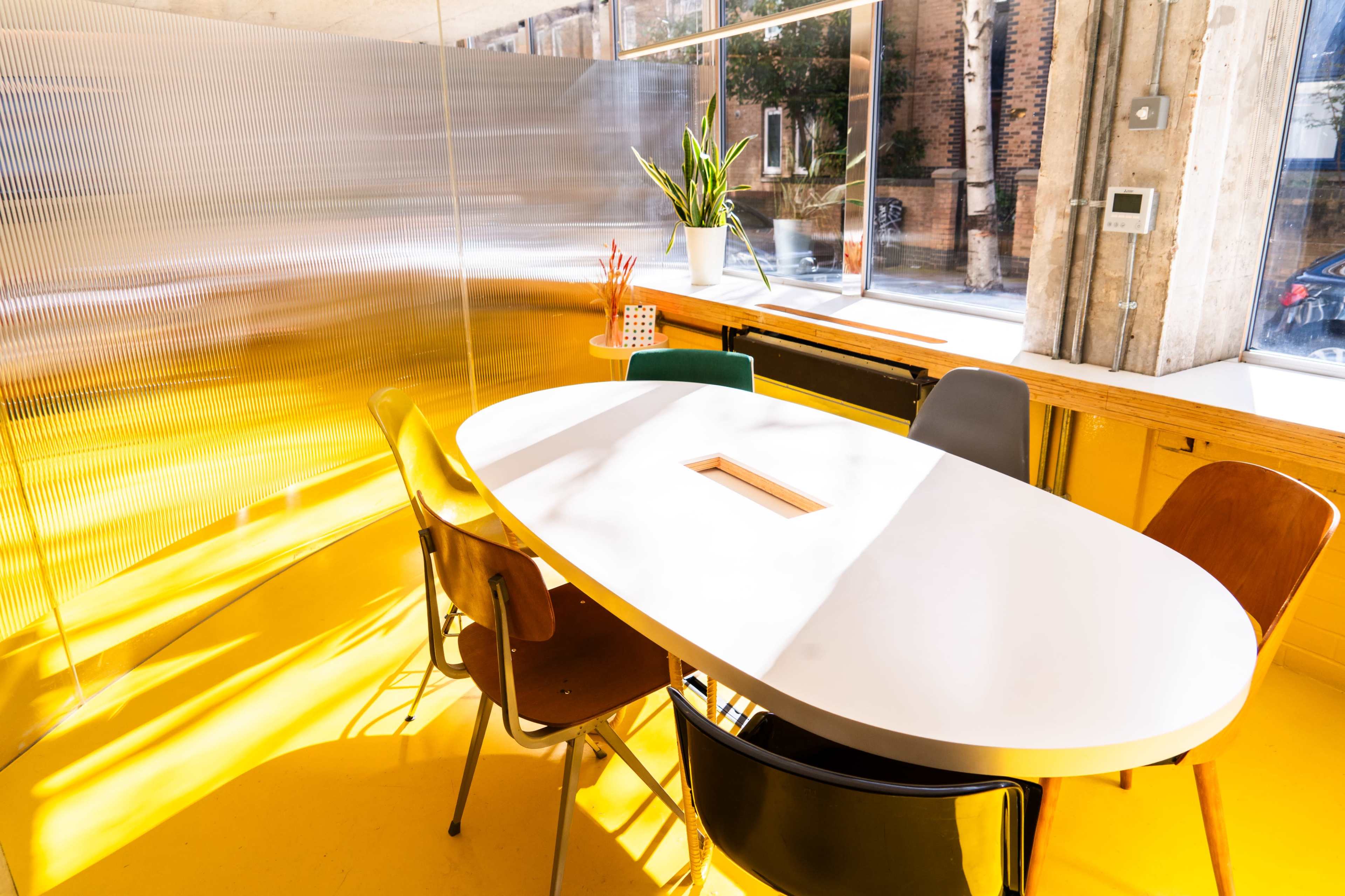 The image shows a brightly lit meeting room with an oval white table surrounded by colorful chairs, against a backdrop of a shiny metallic wall.
