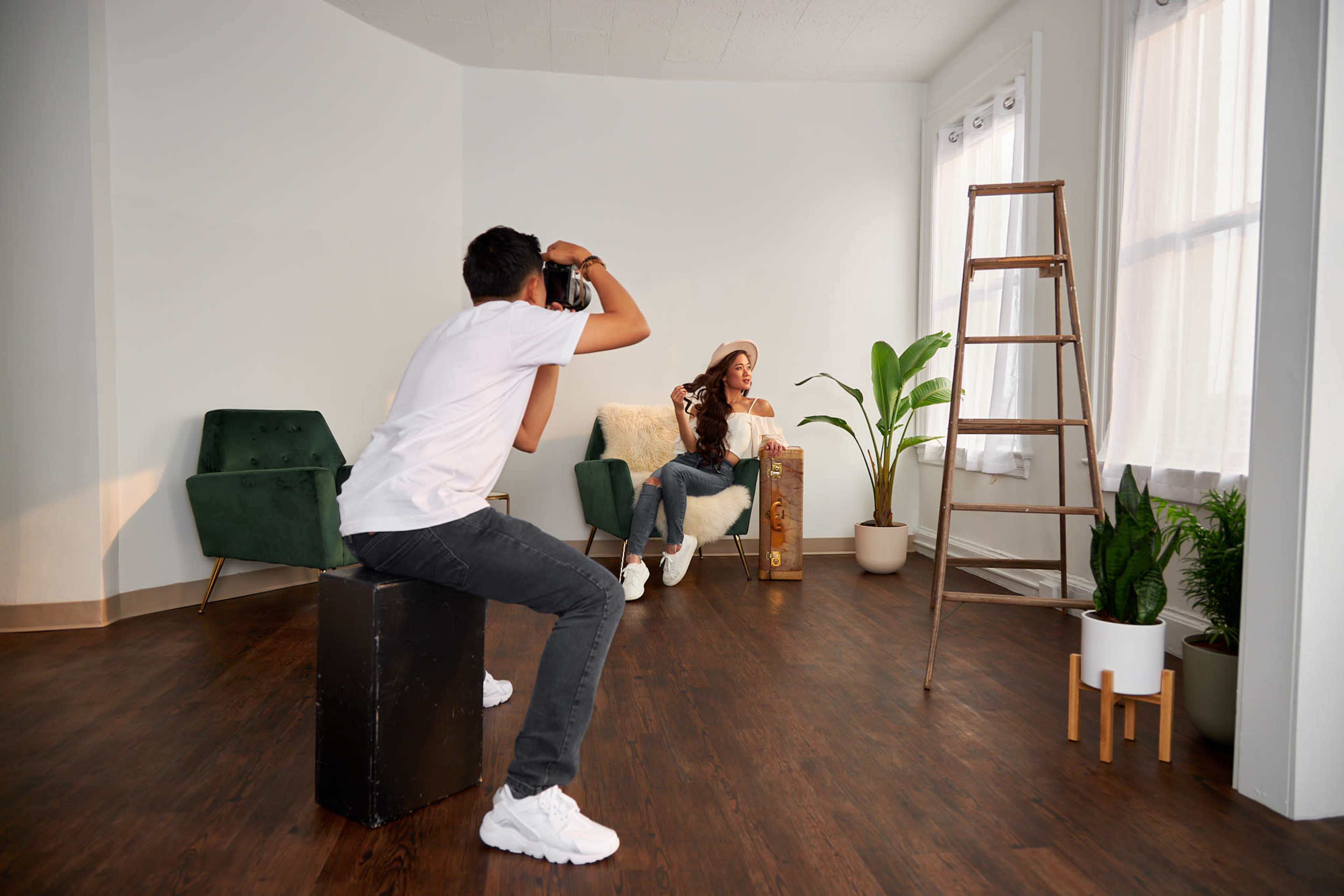 A photographer is capturing an image of a woman sitting on a chair in a well-lit room with plants and furniture.
