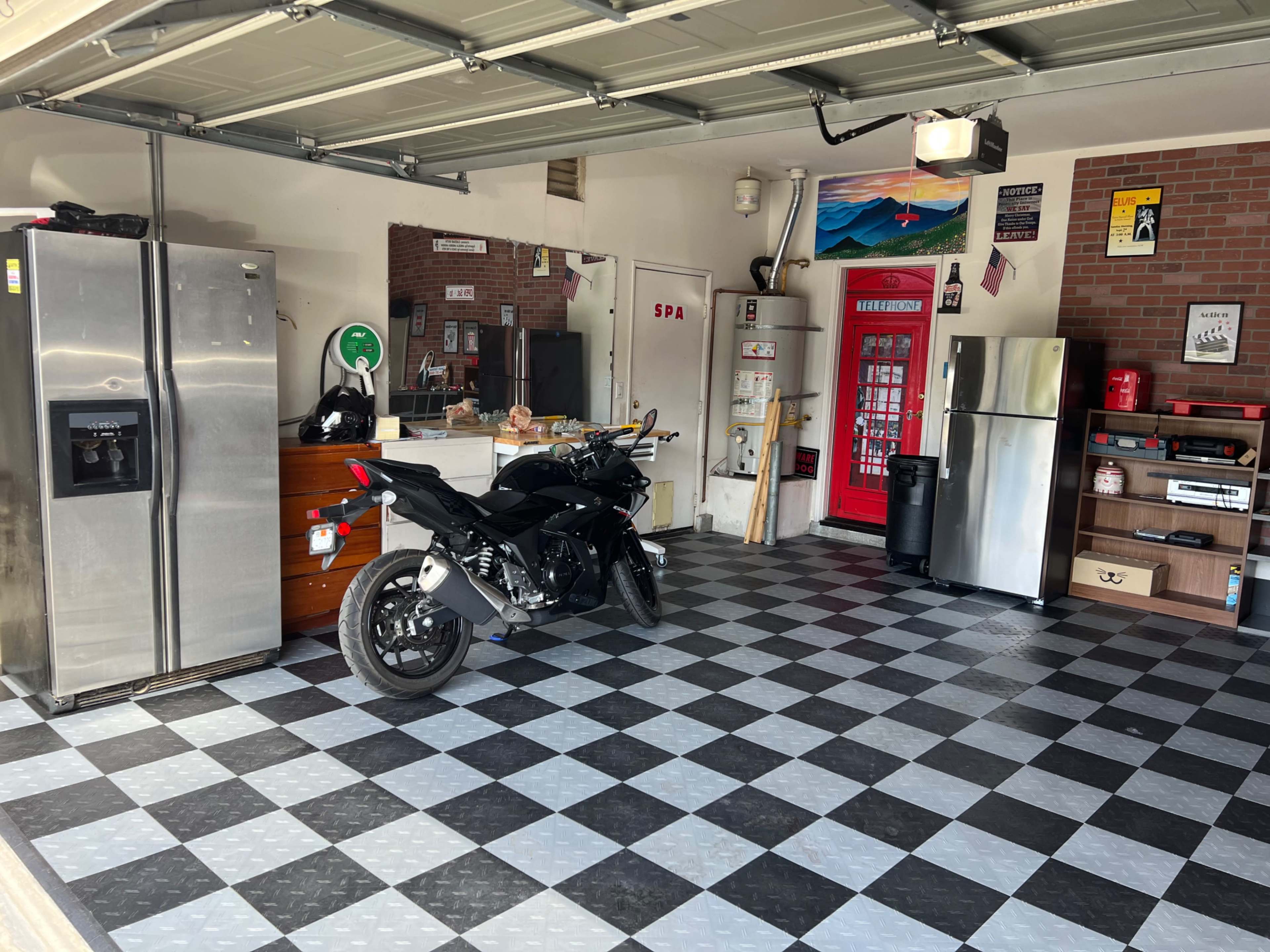 A garage with a motorcycle parked on a black and gray checkered floor, a stainless steel refrigerator, and various items stored on shelves and cabinets against the walls.