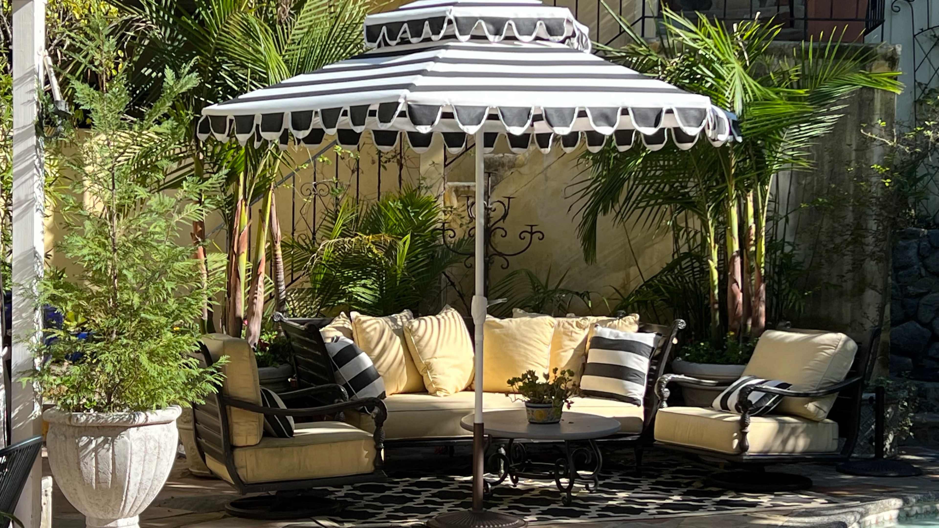 A patio seating area features a round table and cushioned chairs under a large striped umbrella, surrounded by greenery.