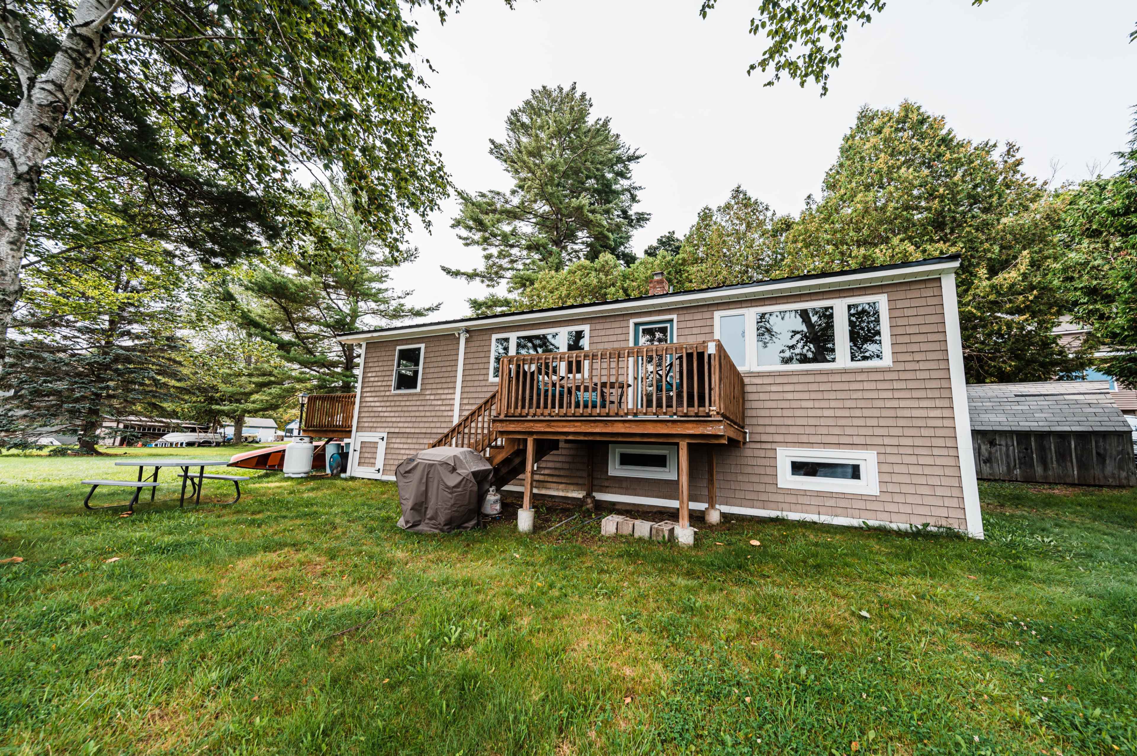 A single-story house with a wooden deck and a picnic table sits in a grassy yard surrounded by trees.