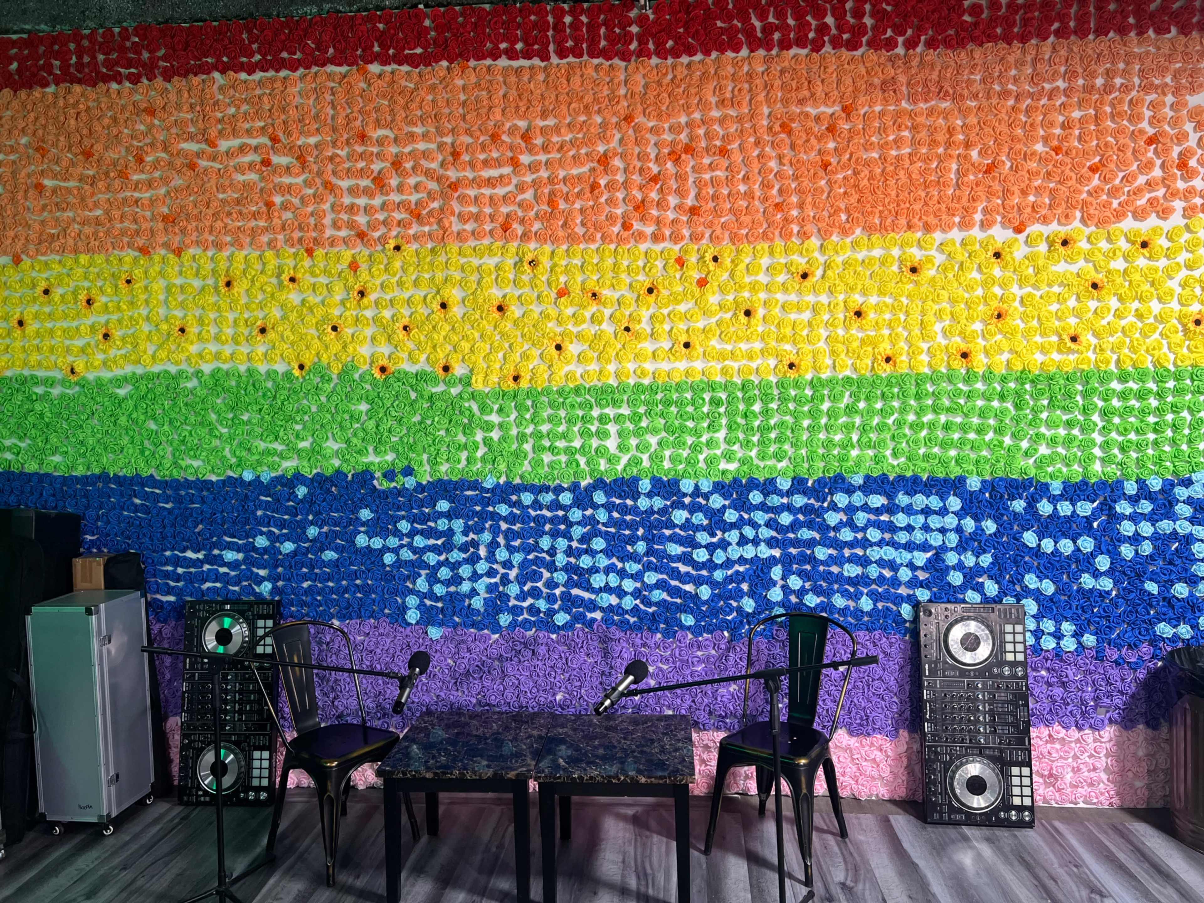 The image shows a colorful backdrop made of fabric or paper flowers arranged in a rainbow pattern, with two microphones and a table in front of it in an indoor setting.