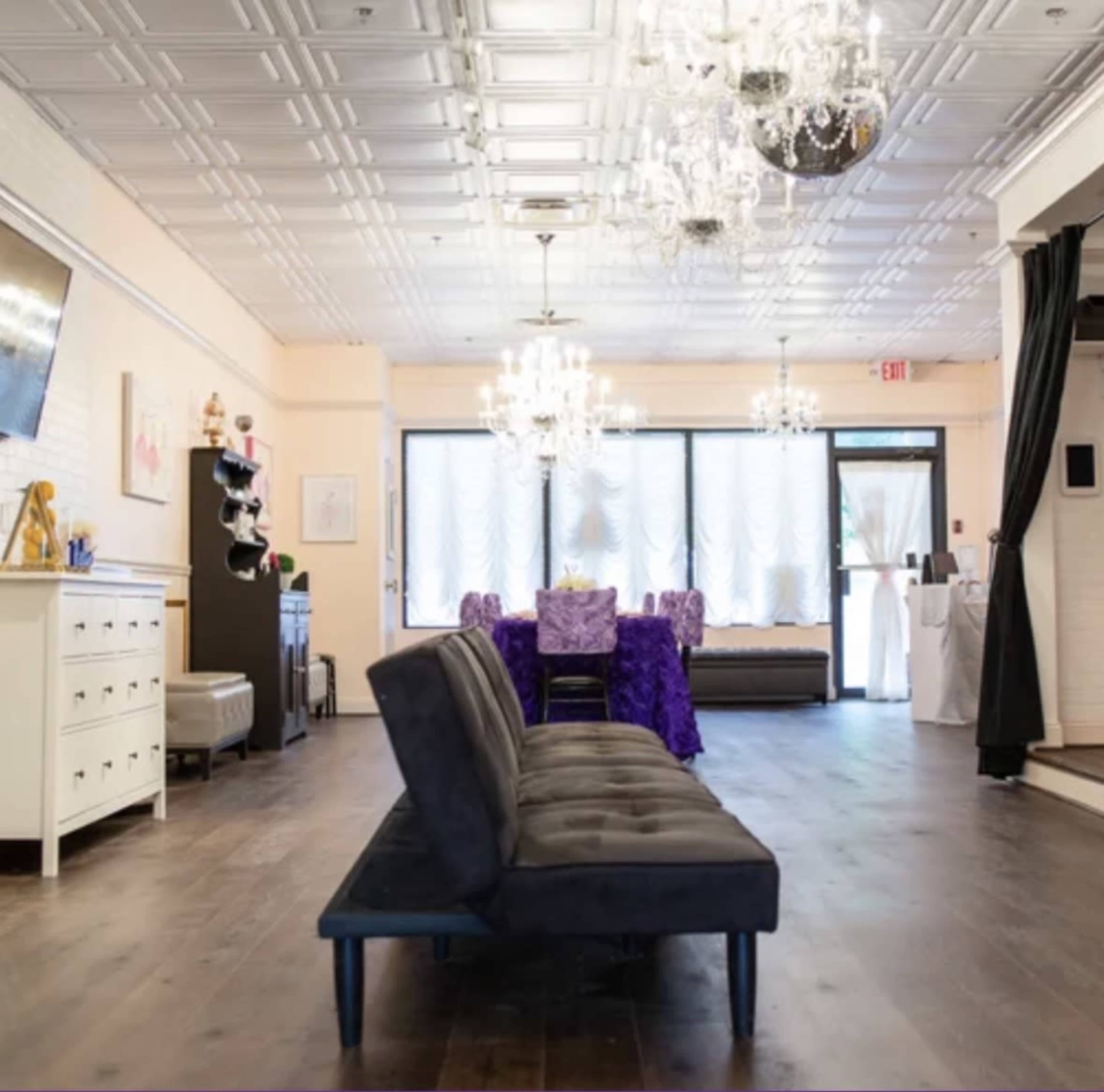 The image shows a spacious, well-lit interior of a modern venue with stylish furniture, chandeliers, and a table covered in purple decor.