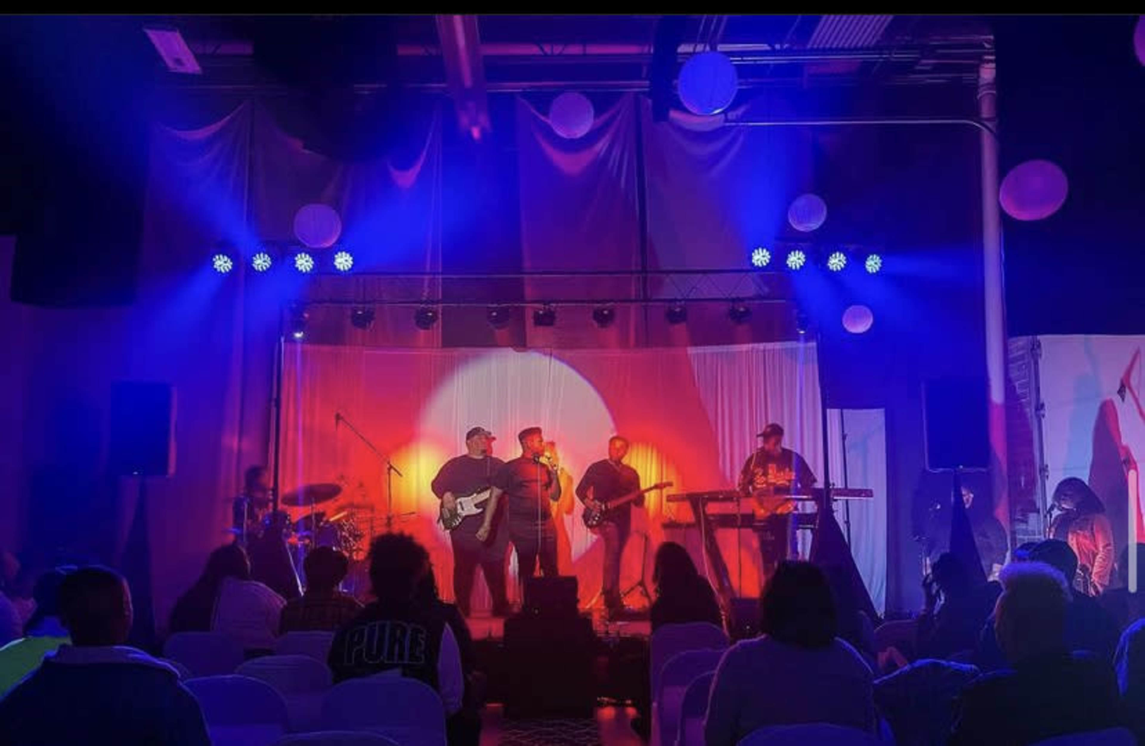 A musical performance takes place on stage with a band and singers, illuminated by vibrant lights in a dimly lit venue.