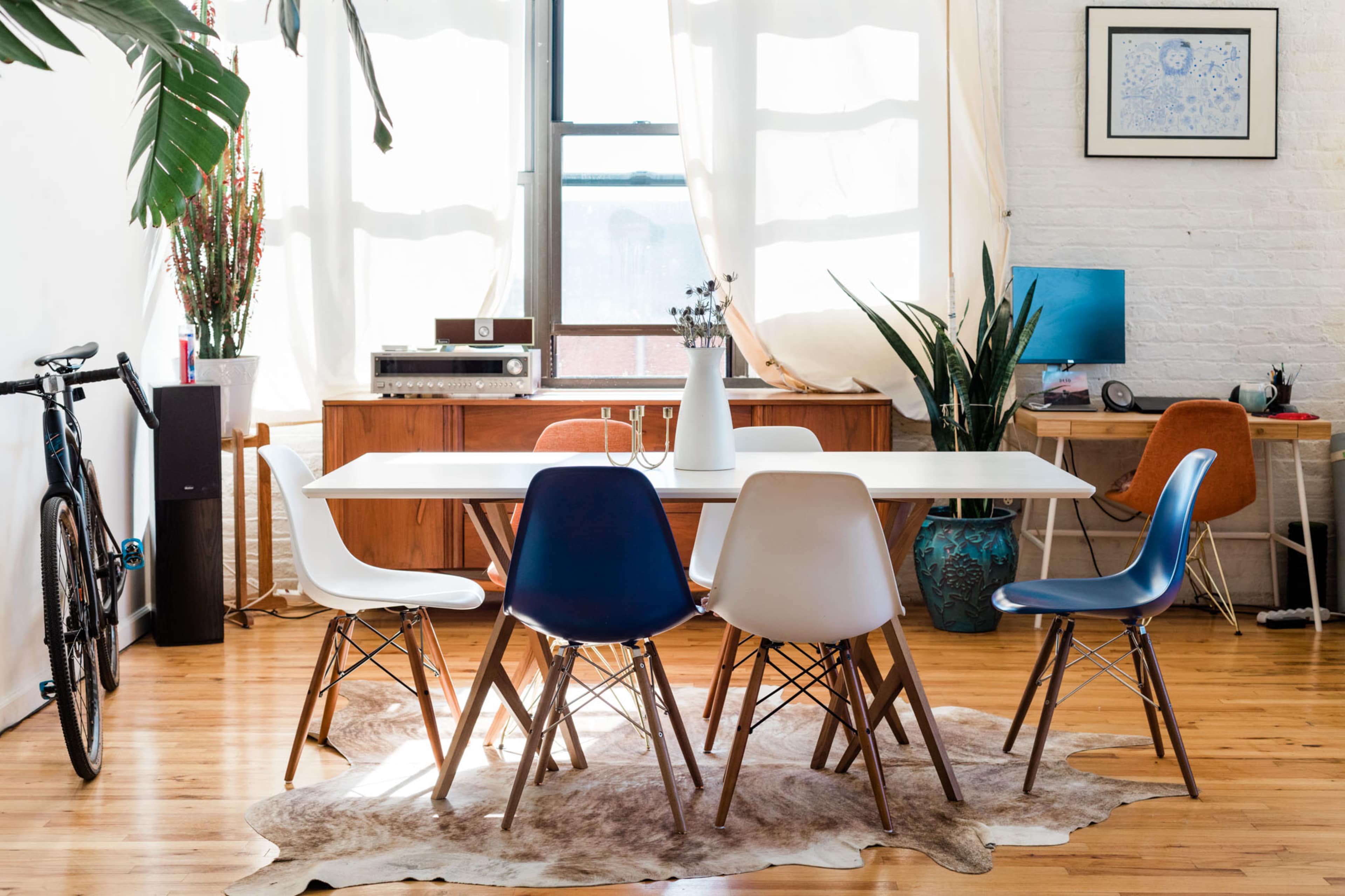 A bright dining area features a white table surrounded by five colorful chairs, with a wooden sideboard, plants, and a desk in the background.