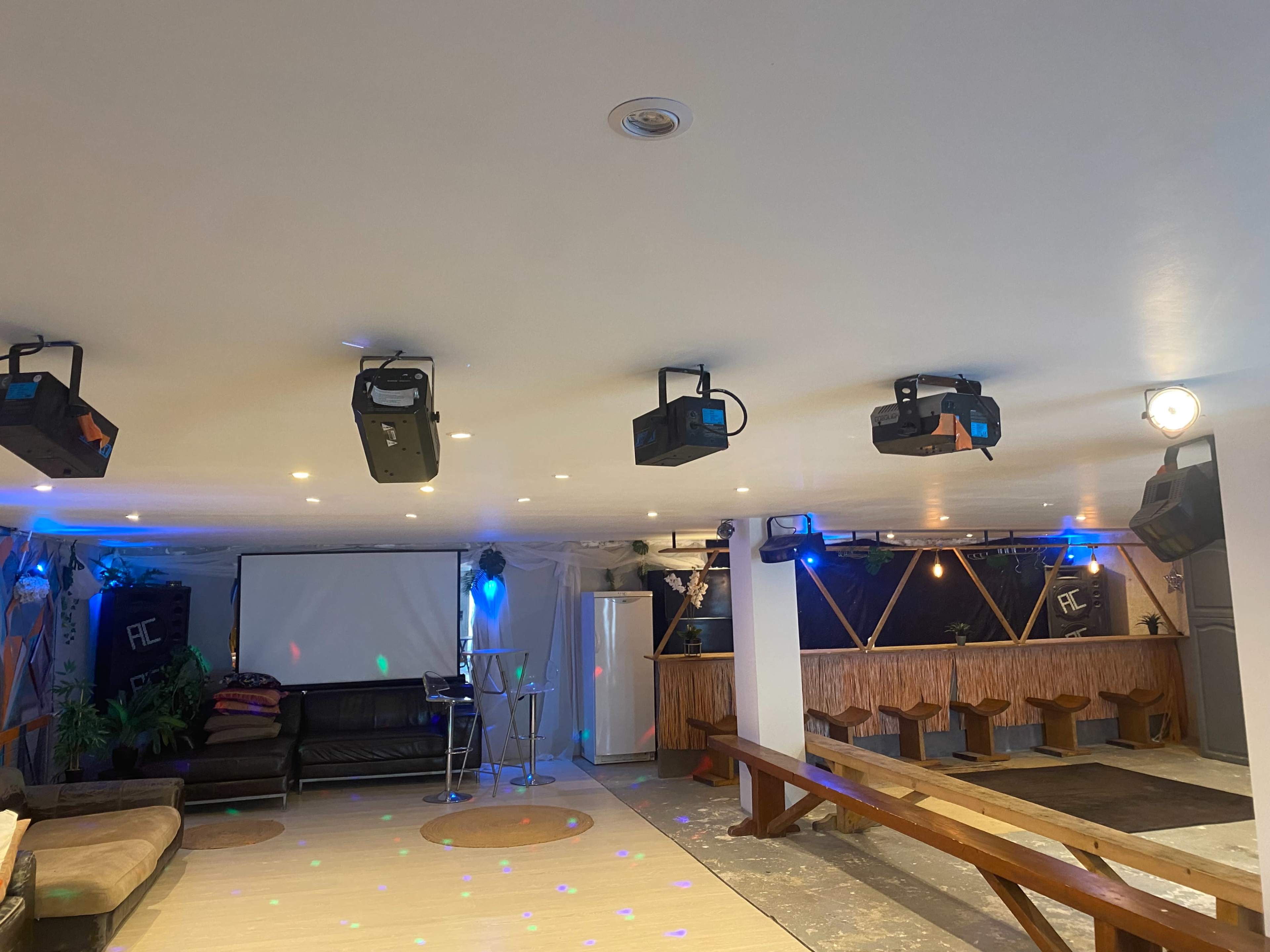 A spacious indoor venue featuring a seating area with a couch, a screen at the front, and a bar setup with colorful lighting and projection equipment.