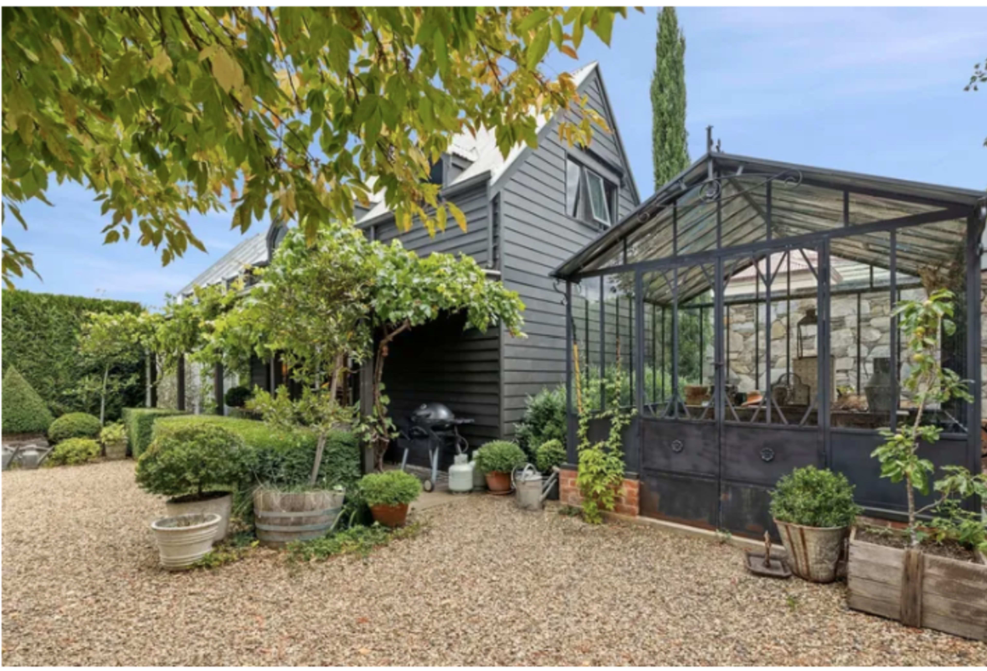 The image shows a landscaped garden with a gravel path leading to a black wooden house and a glass greenhouse surrounded by various potted plants and trimmed hedges.