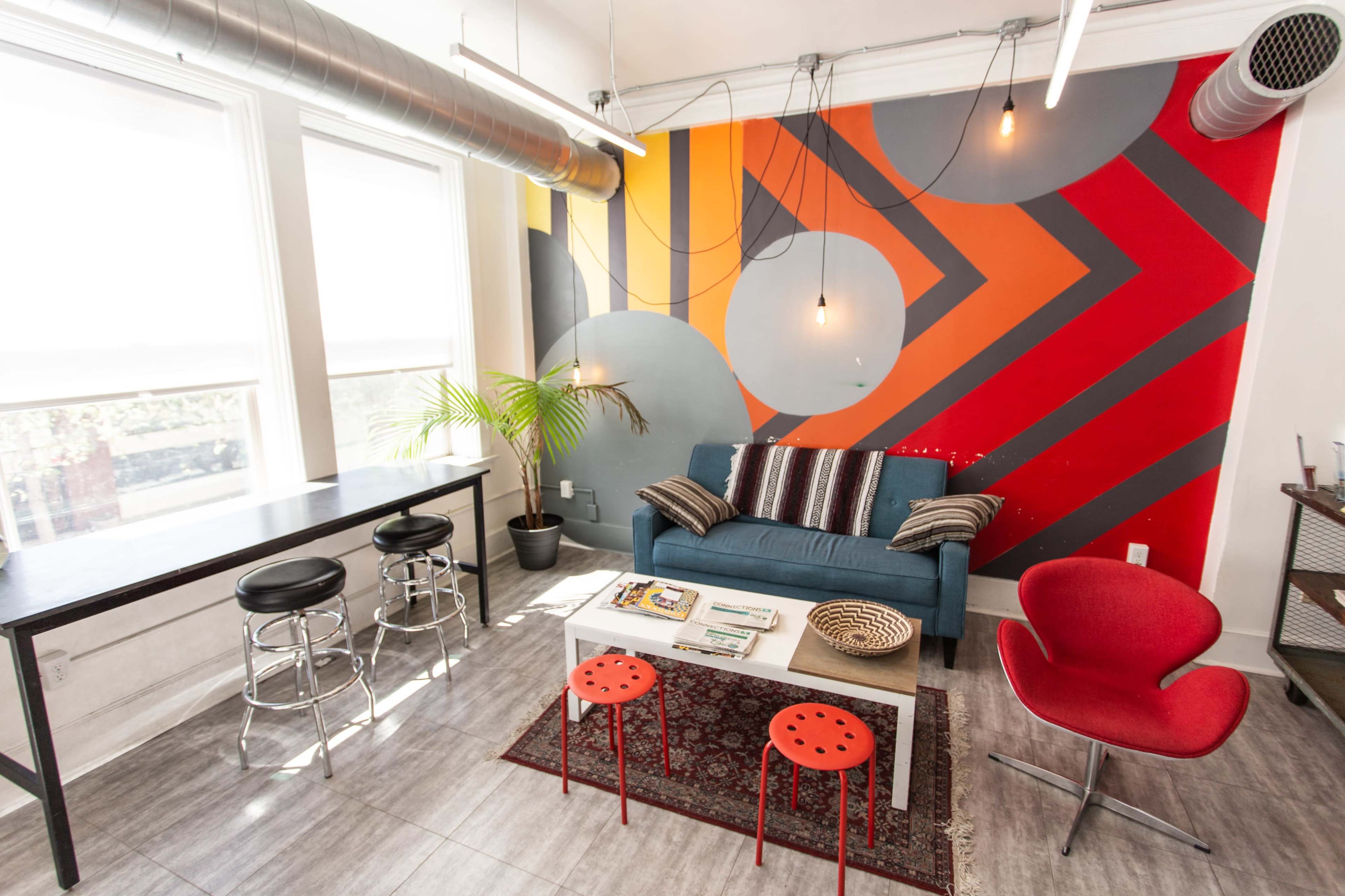 A brightly designed living space featuring a blue sofa, a white coffee table with red stools, and a mural of geometric shapes on the wall.