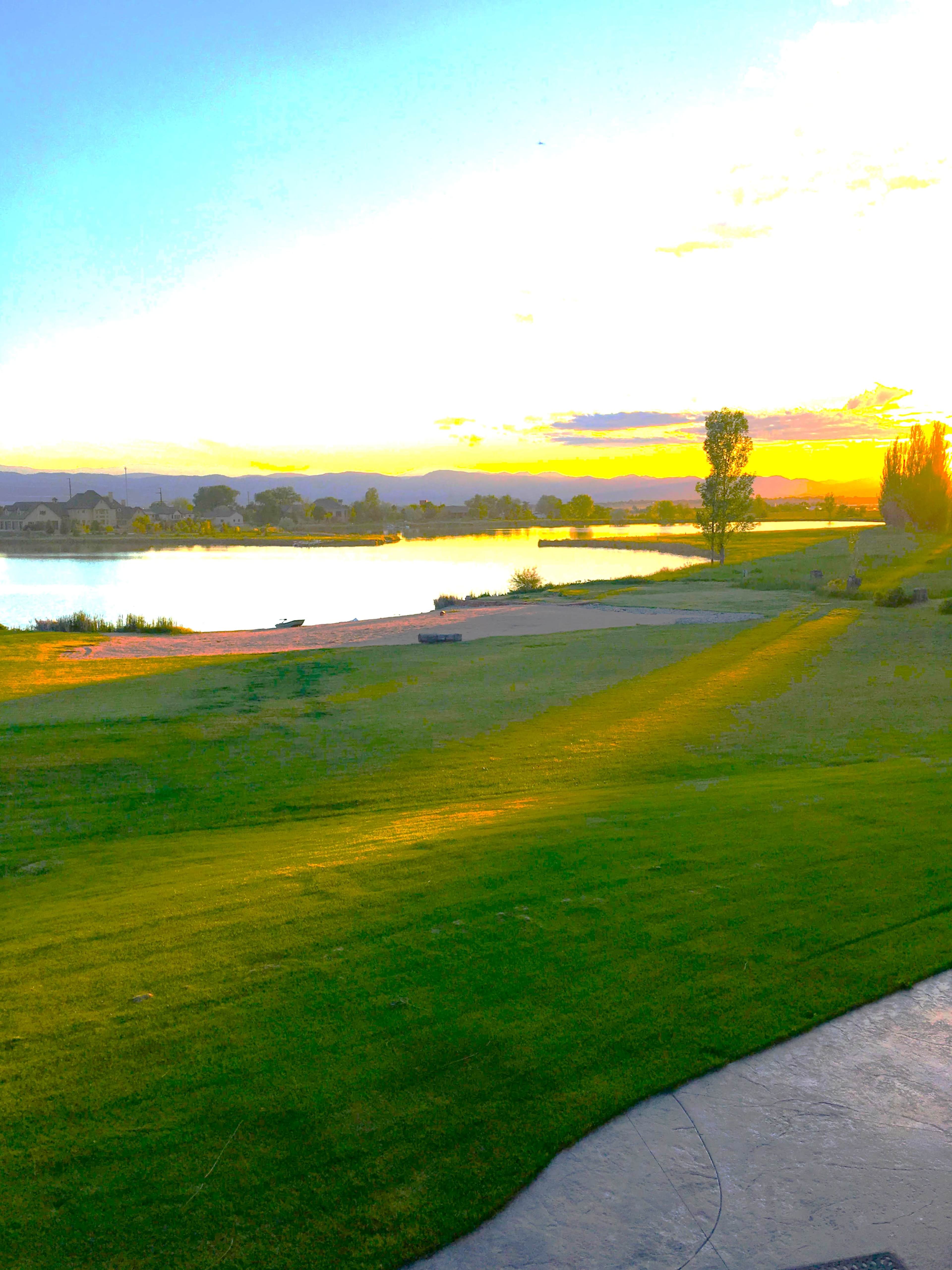 A grassy area slopes down to a calm lake, with a sunset casting a warm glow across the landscape.