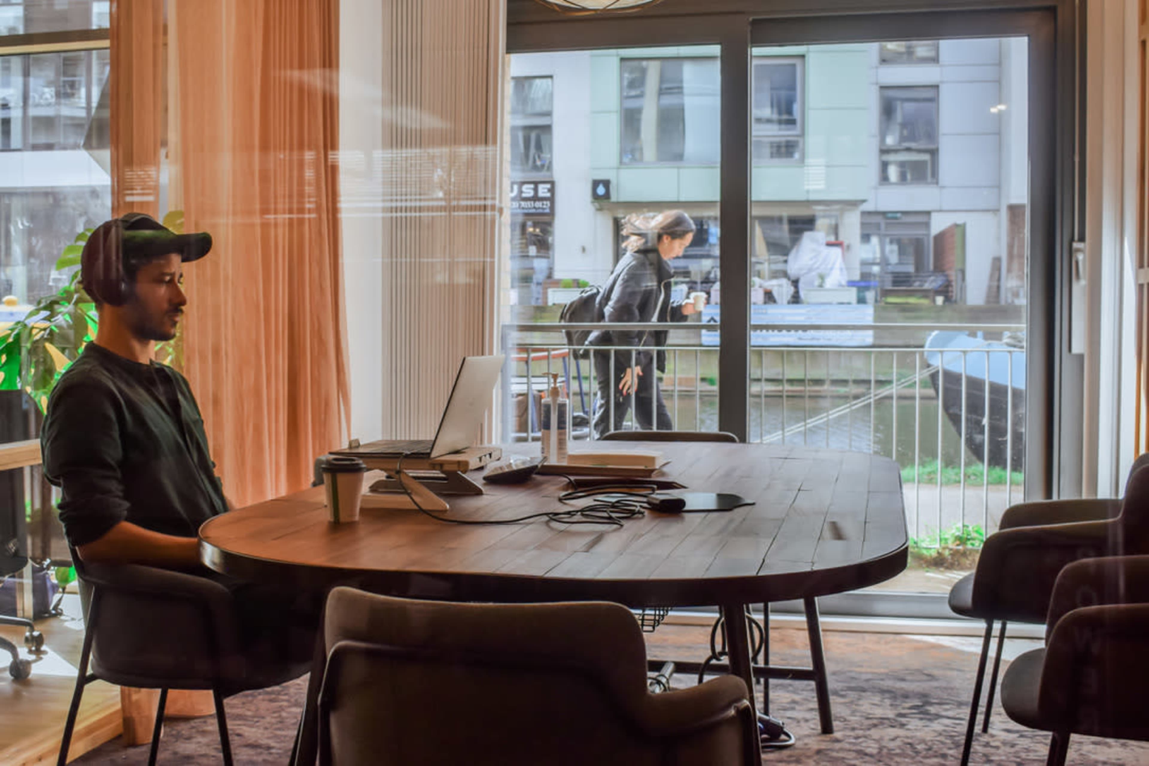 A man sits at a round wooden table with a laptop and coffee cup, while a woman walks by a large window overlooking a canal.