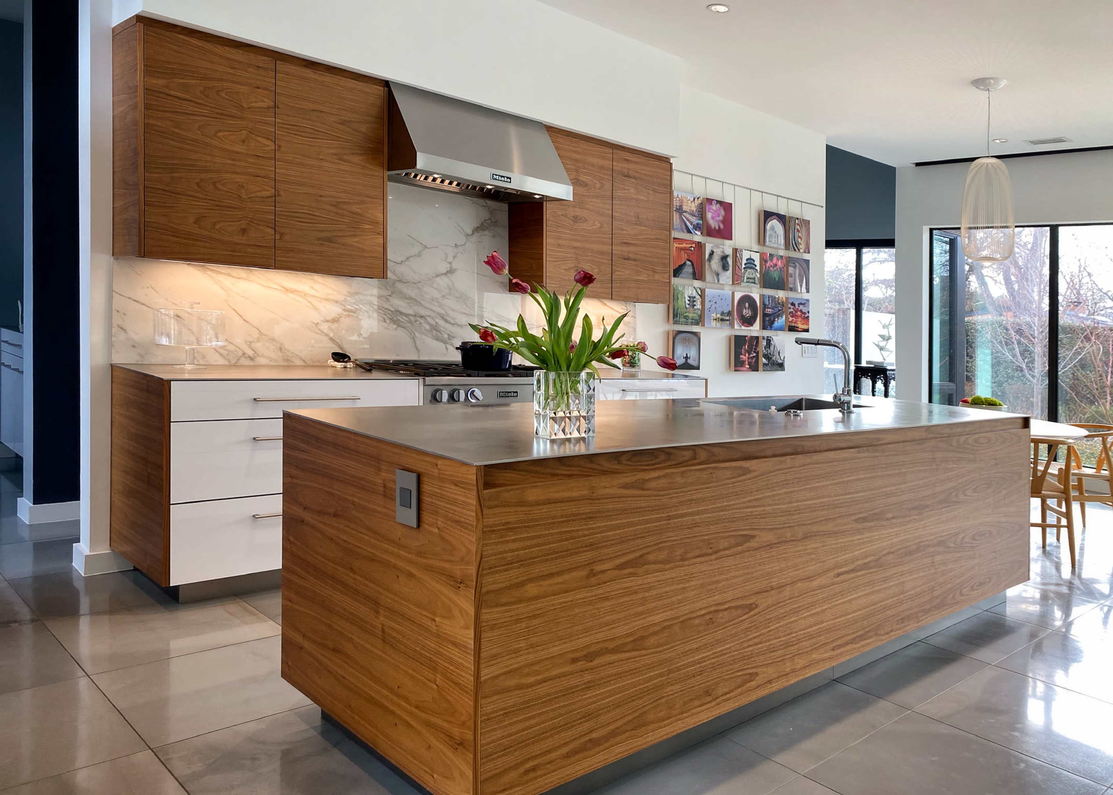 The image shows a modern kitchen with wood-paneled cabinetry, a marble backsplash, a stainless steel range hood, and a large island with a vase of tulips on it.