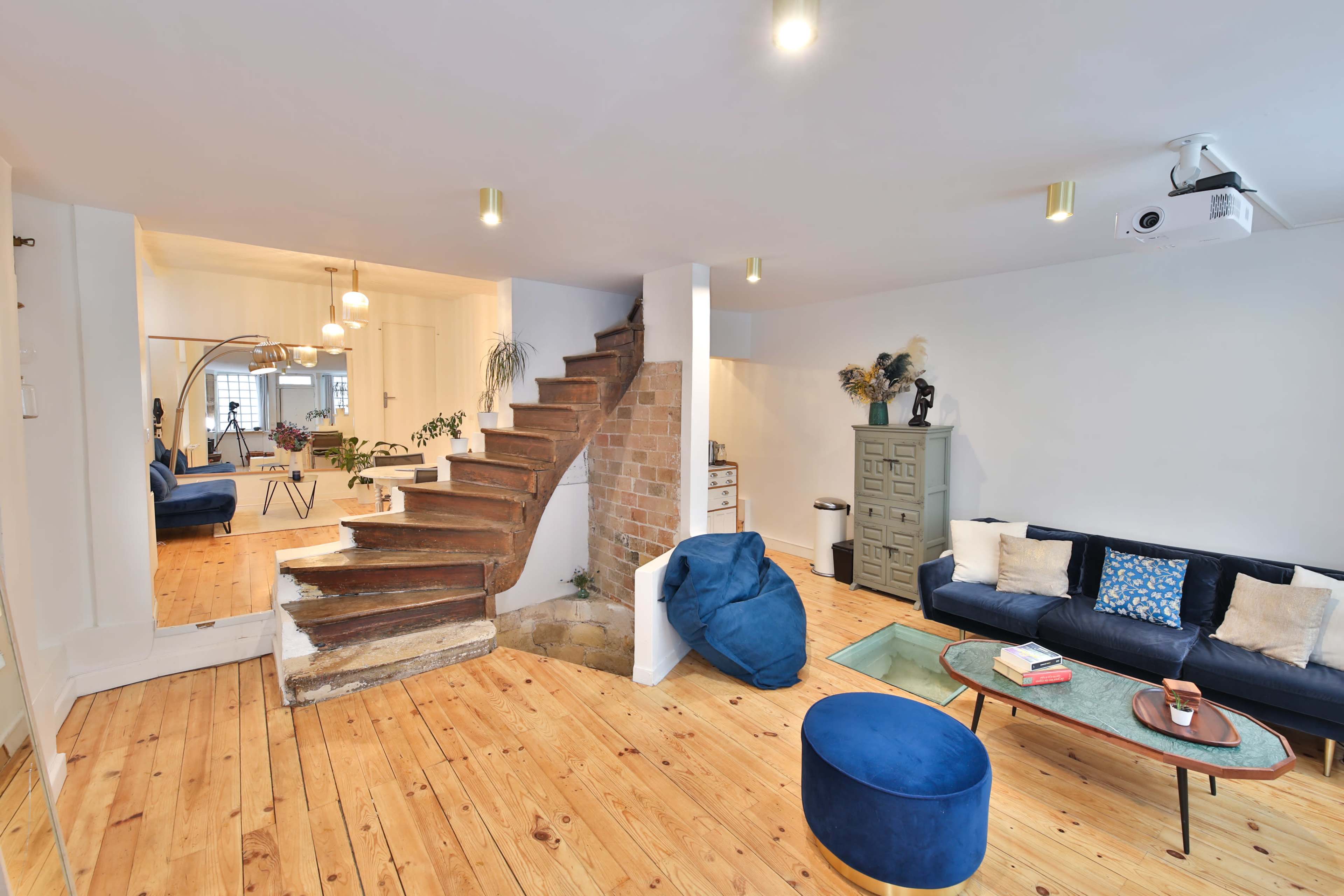 A modern living space with a wooden staircase, a blue sofa, a circular ottoman, and a projector mounted on the ceiling.