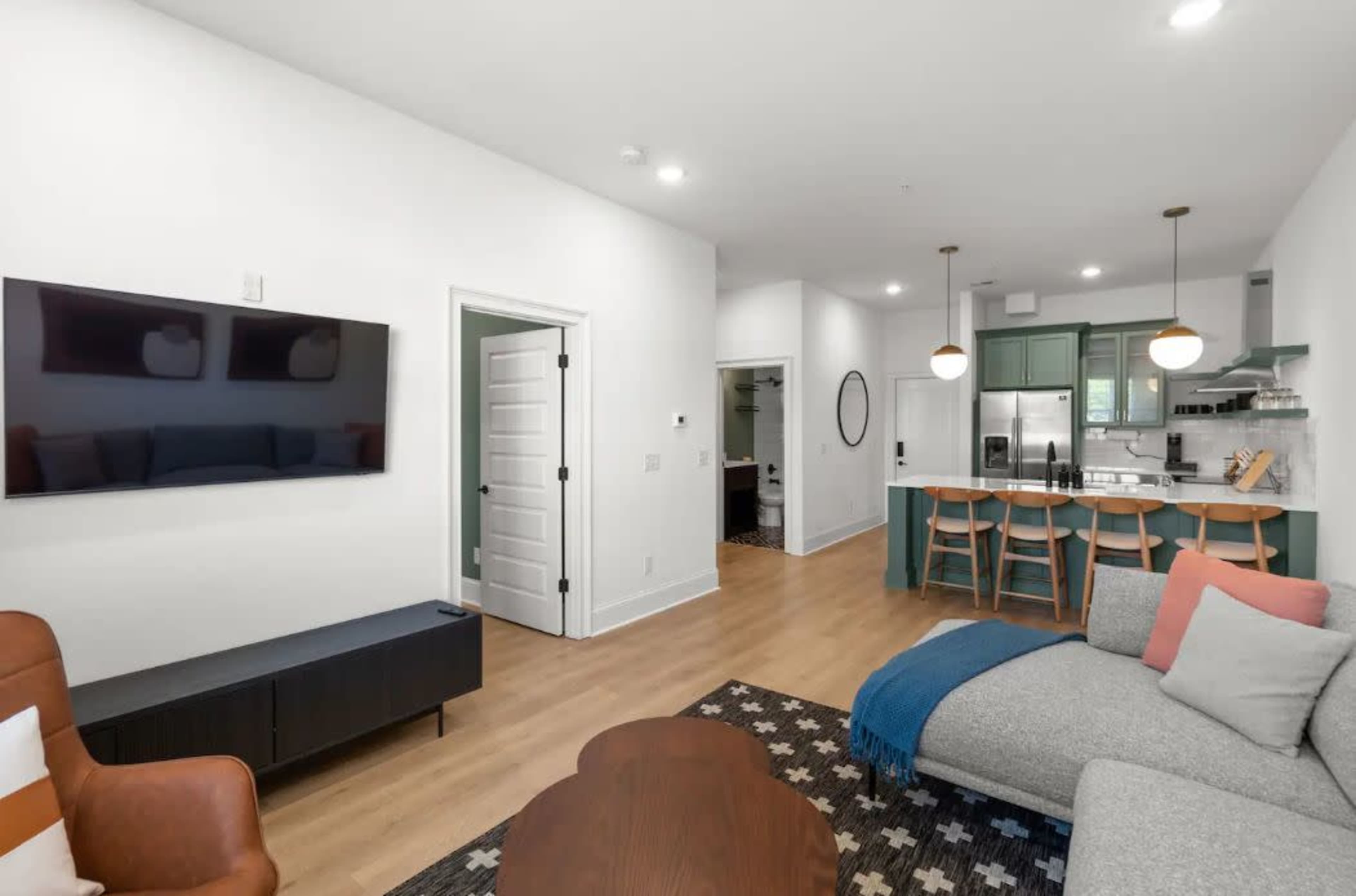 A modern living room and kitchen area featuring a flat-screen TV, a brown armchair, a gray sofa, and a kitchen with green cabinets and bar seating.