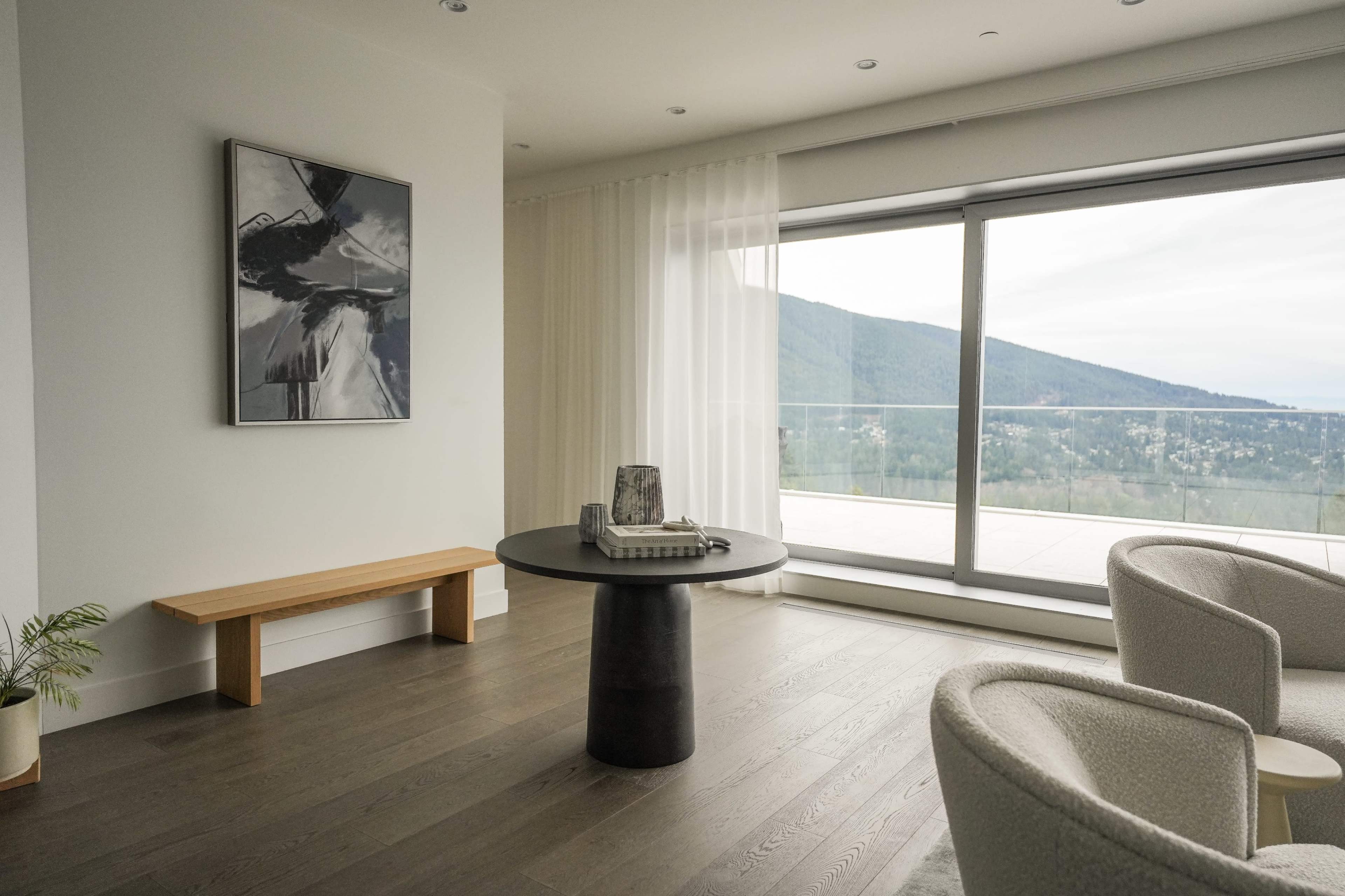 A modern, minimalist living room with a small round table, two light-colored chairs, a wooden bench, and large windows overlooking a mountain view.