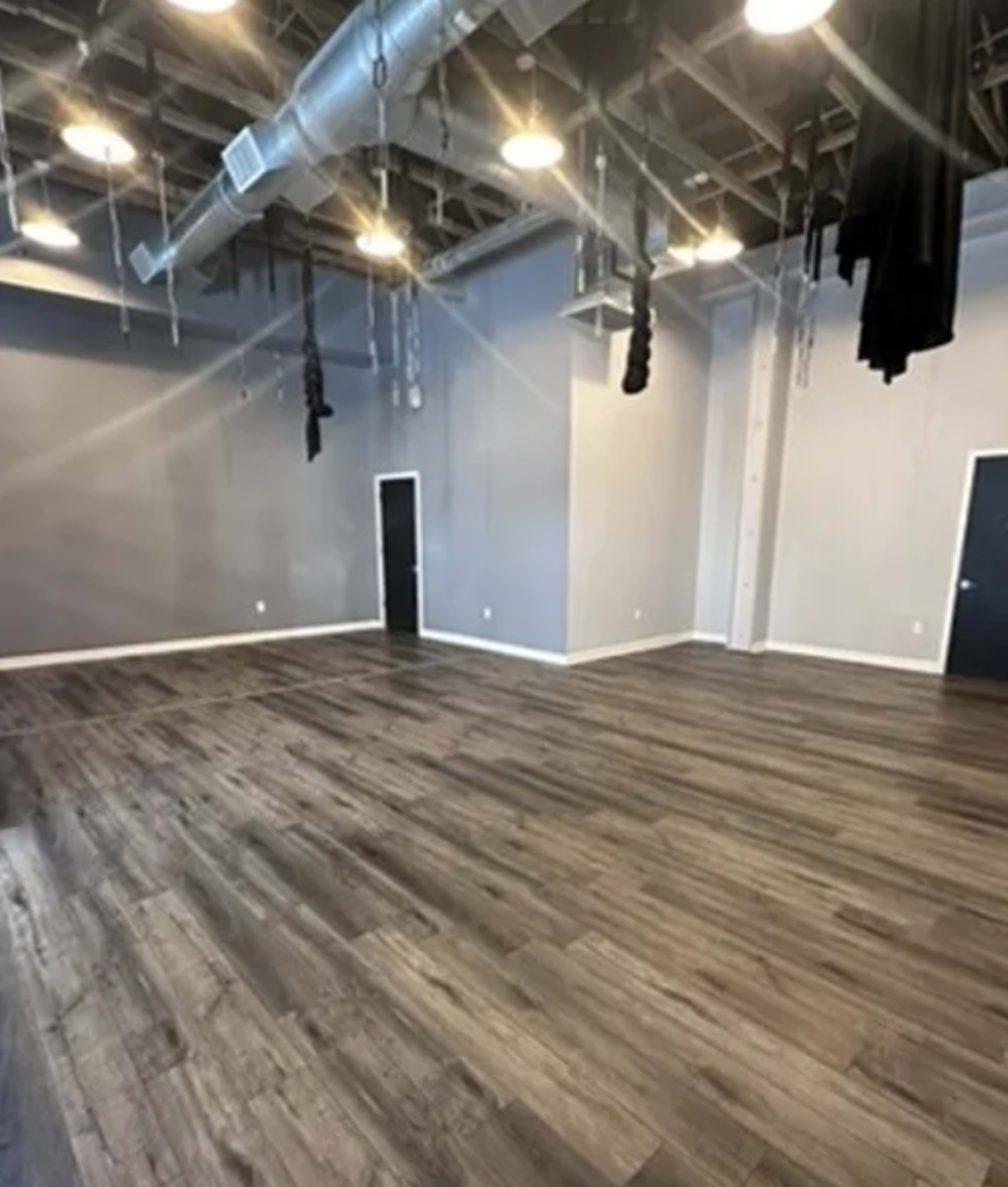 A spacious room with a hardwood floor, high ceilings, and dark wall panels, featuring two doors on one side and hanging fabric or ropes from the ceiling.