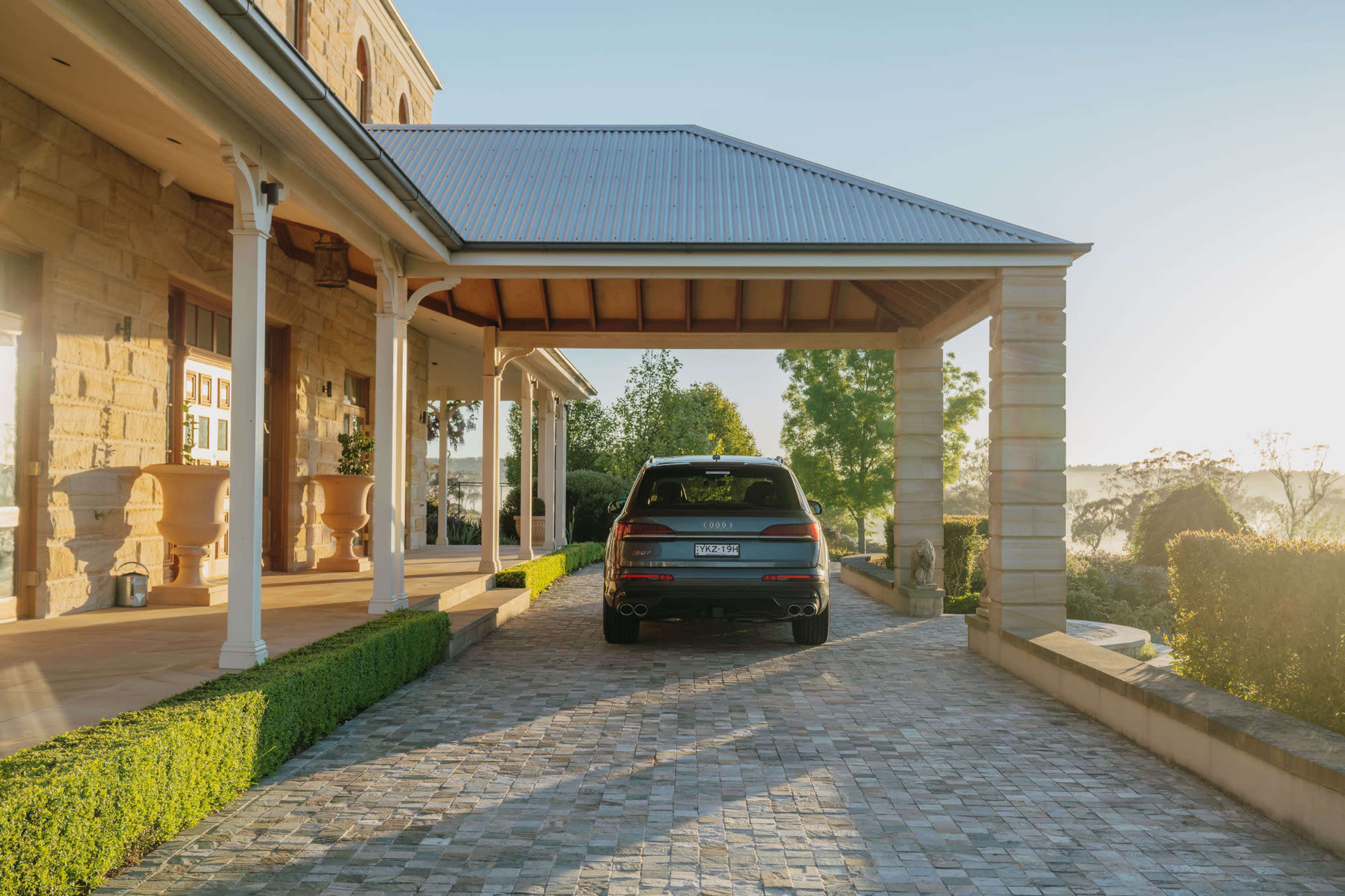 A car is parked under a covered area of a stone house, with a landscaped driveway leading to it.
