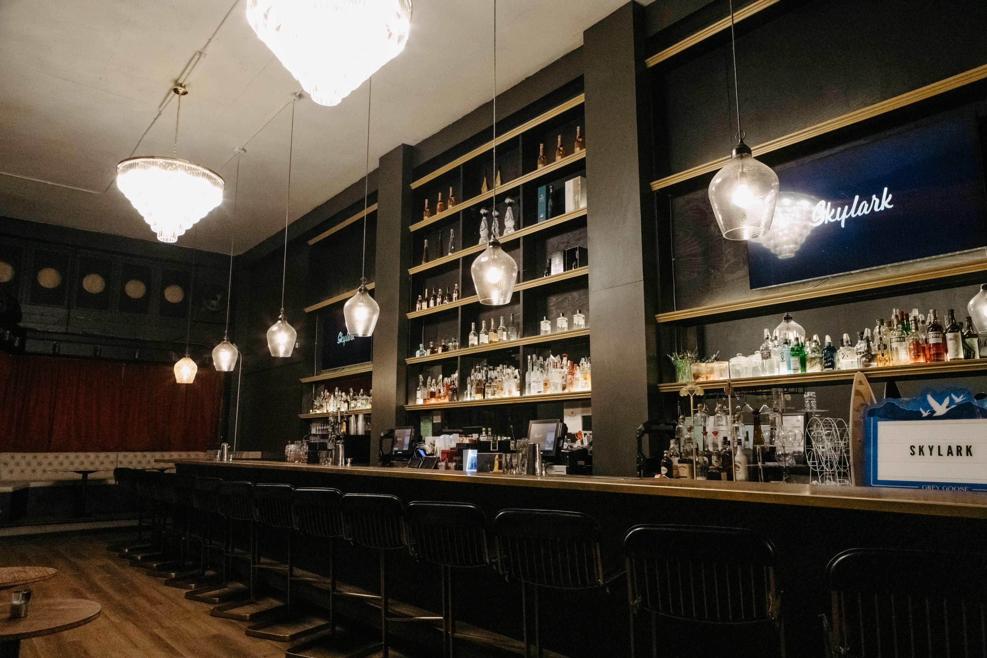 The image shows a well-lit bar area with shelves filled with various bottles and decorative lighting fixtures overhead.