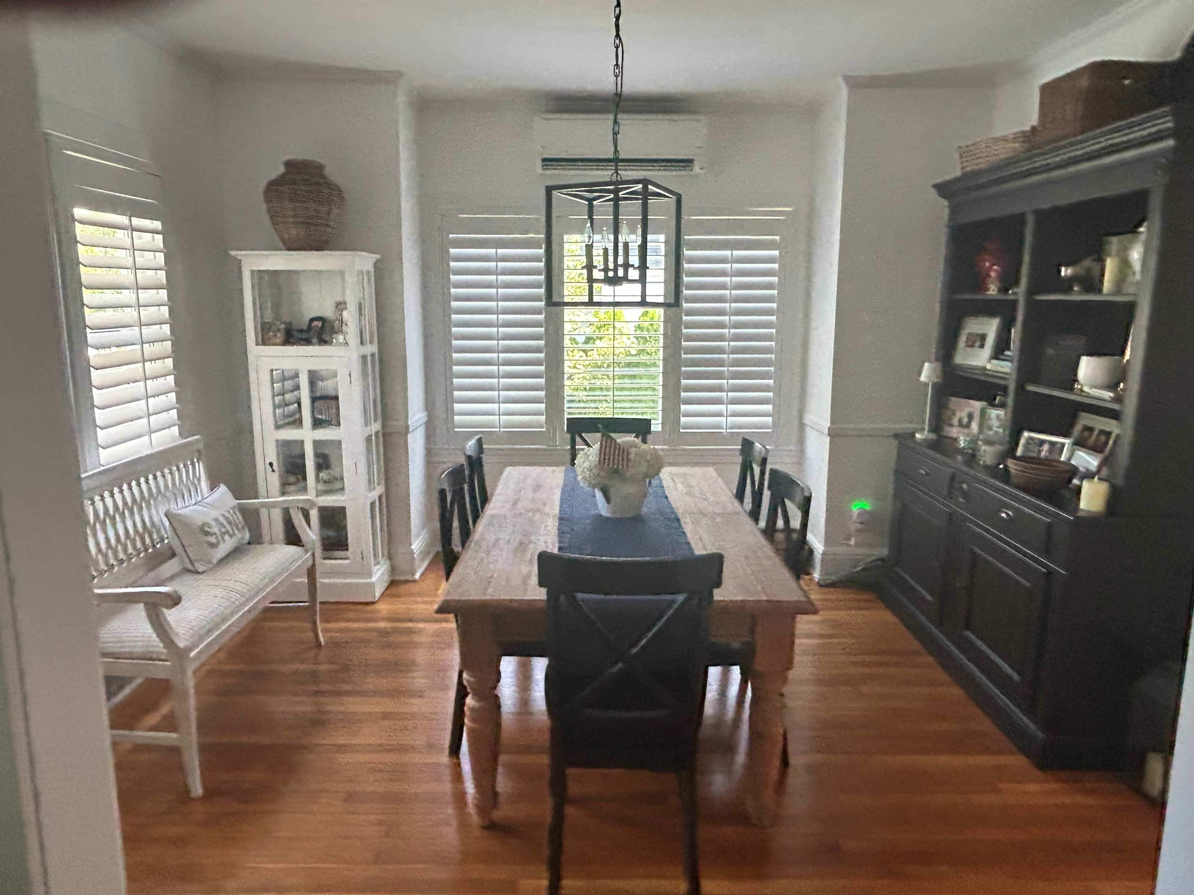 A dining room features a large wooden table surrounded by black chairs, with a white bench, a glass display cabinet, and a dark wooden hutch against the walls.