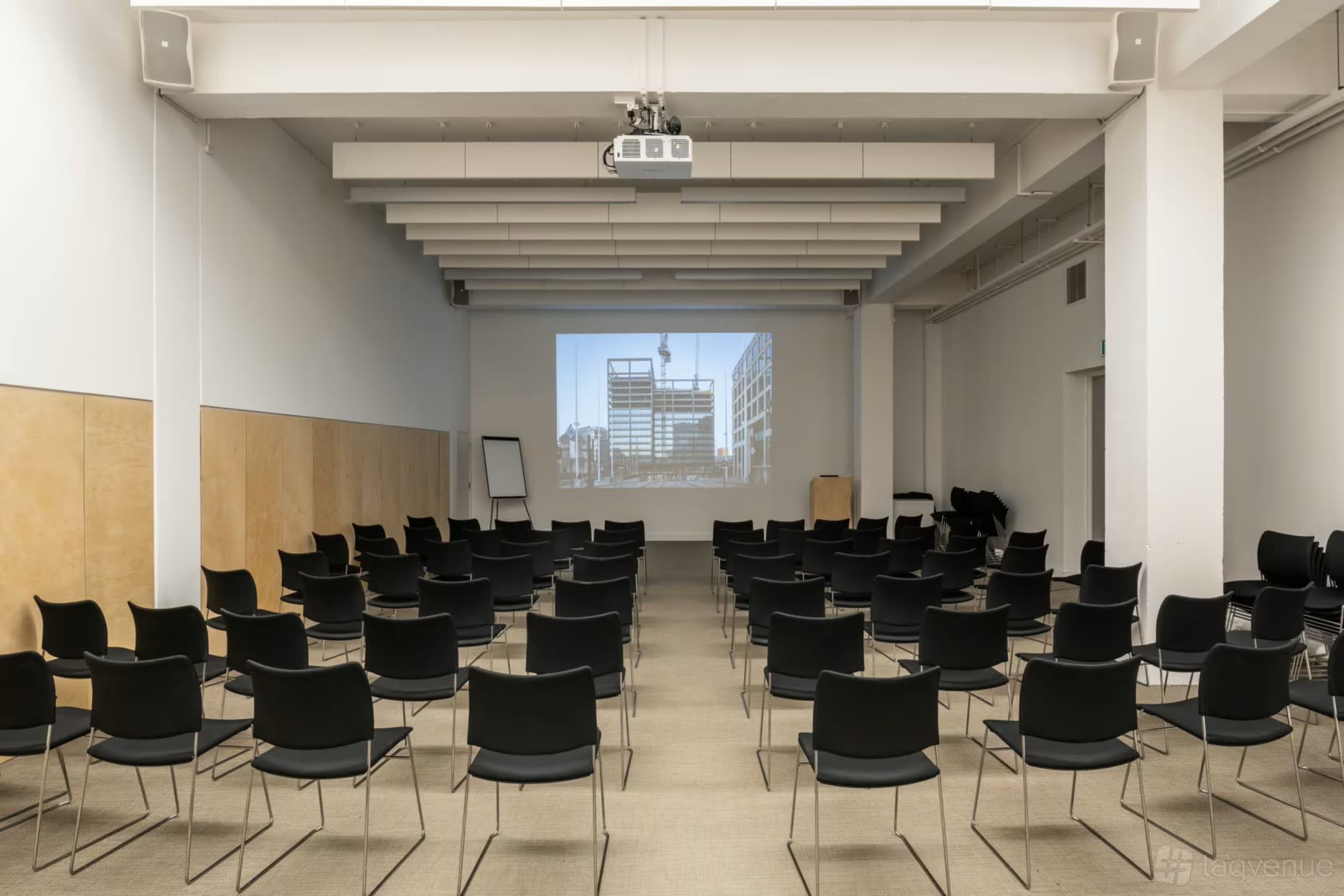 The image shows a spacious conference room arranged with multiple black chairs facing a projection screen displaying a building construction site.