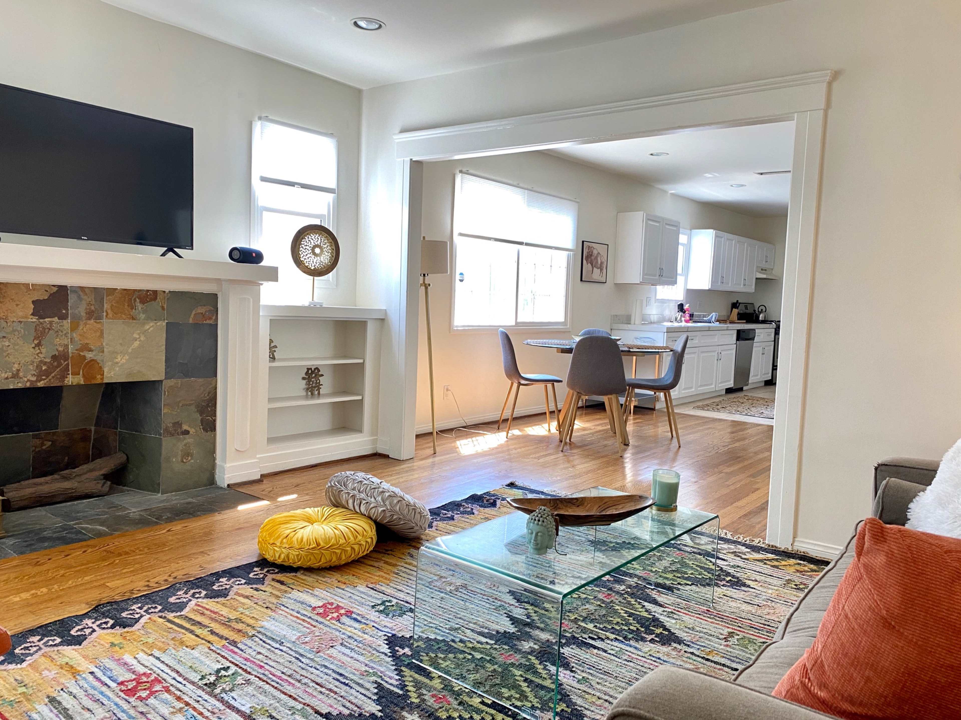 A living room features a television mounted on a white wall, a stone fireplace, and a glass coffee table, with a view into a nearby kitchen.
