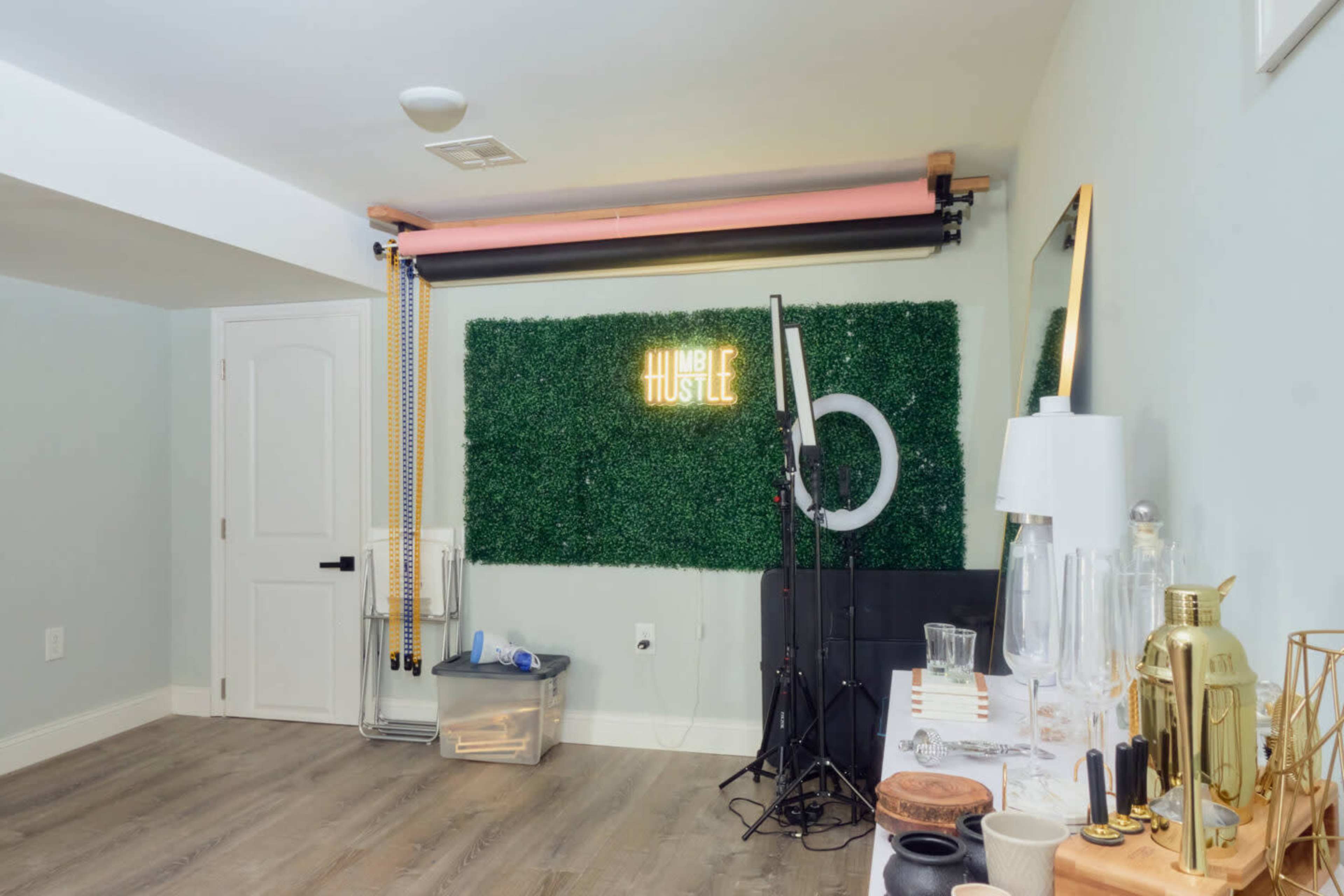 A well-lit room with a green artificial grass wall, a neon sign that reads "HUMBLE HUSTLE," and various photography equipment and decor items arranged on a table.