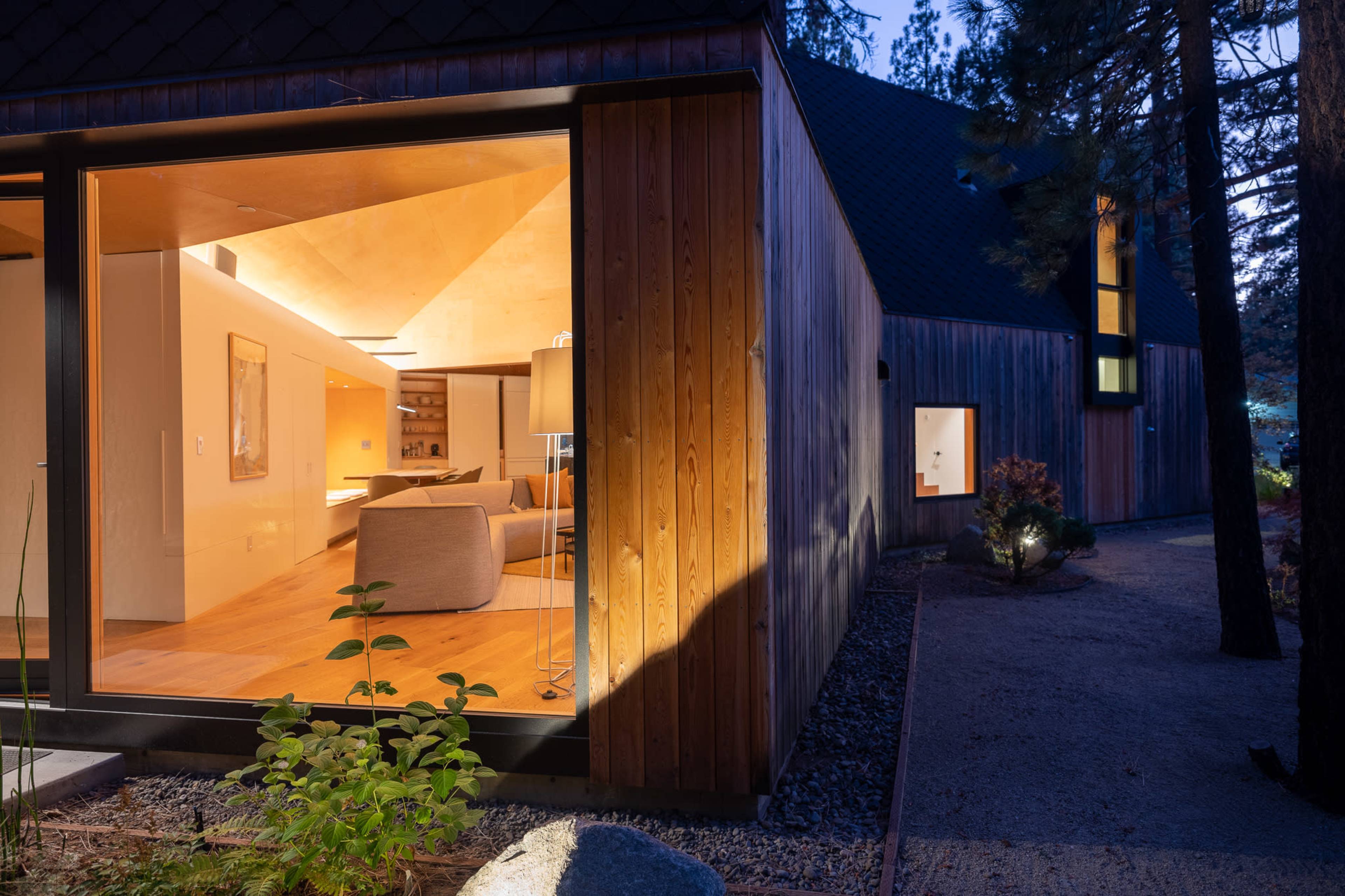 A modern house at dusk features a wooden exterior, large windows, and illuminated interior spaces that showcase a cozy living area.