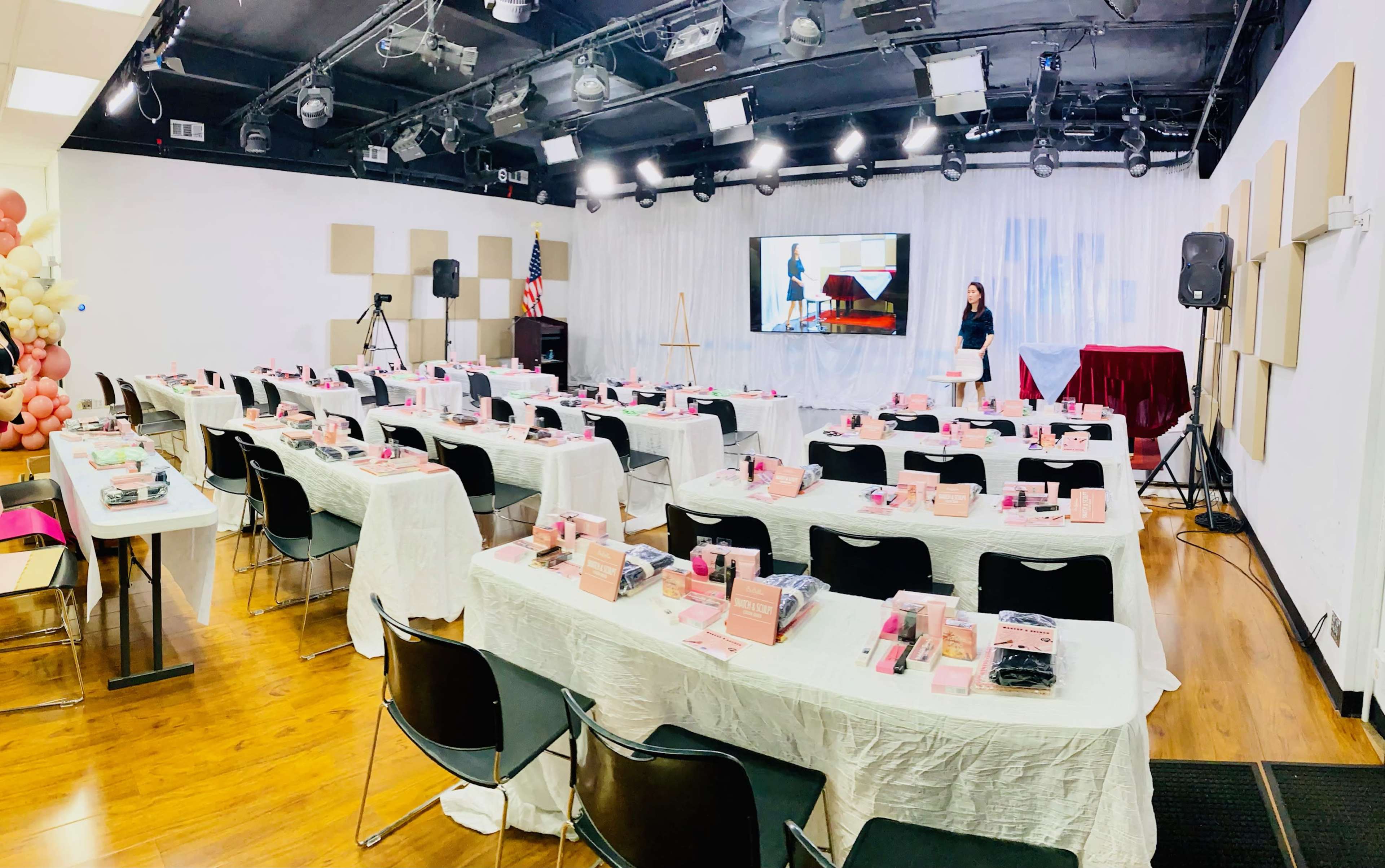 A conference room is set up with multiple tables covered in white tablecloths, each featuring pink presentation materials, and a projector screen displaying a presentation, while a presenter stands in front.