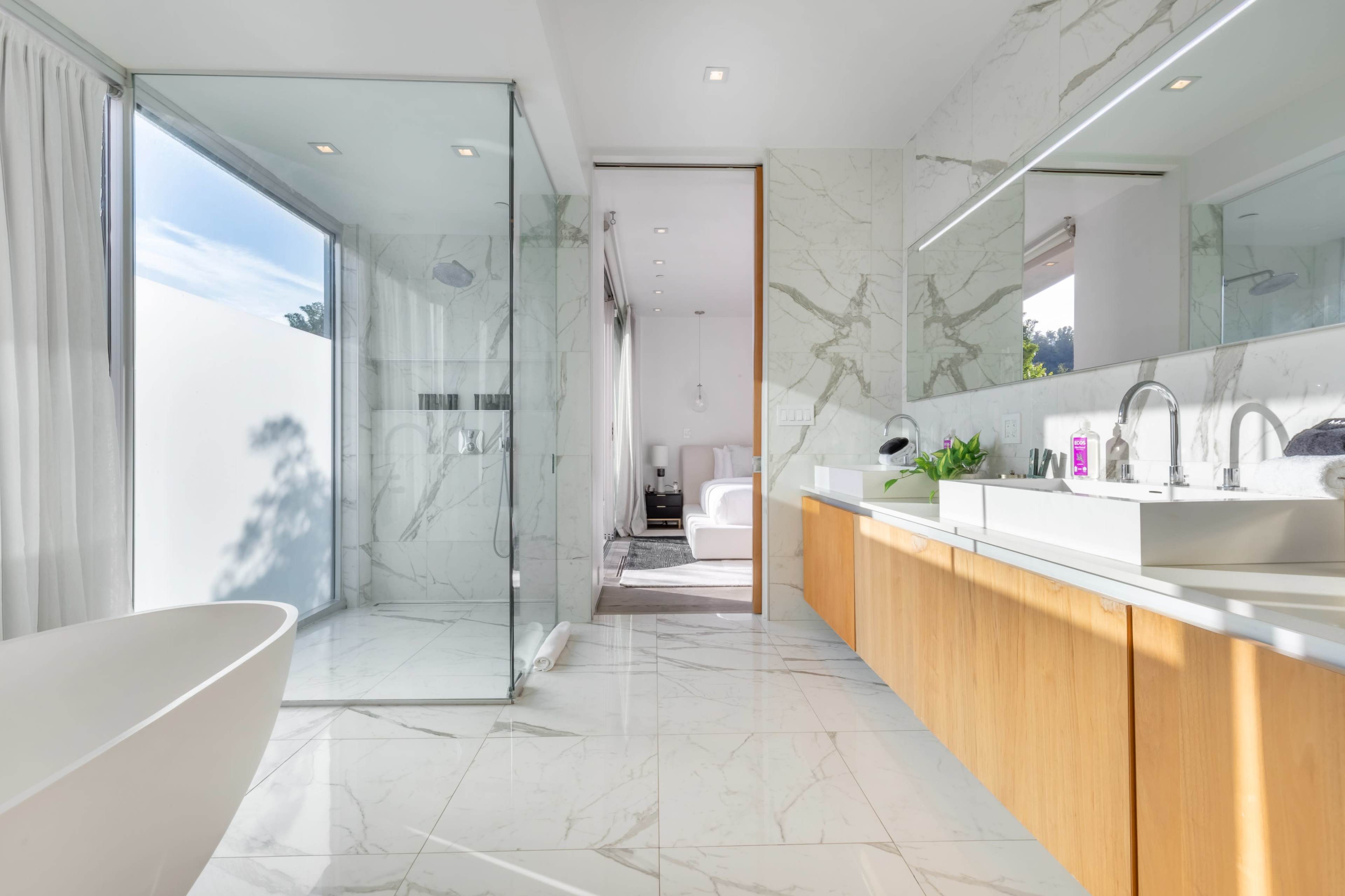 The image shows a modern bathroom with a glass shower, a freestanding bathtub, a double vanity with a wooden cabinet, and large windows providing natural light.