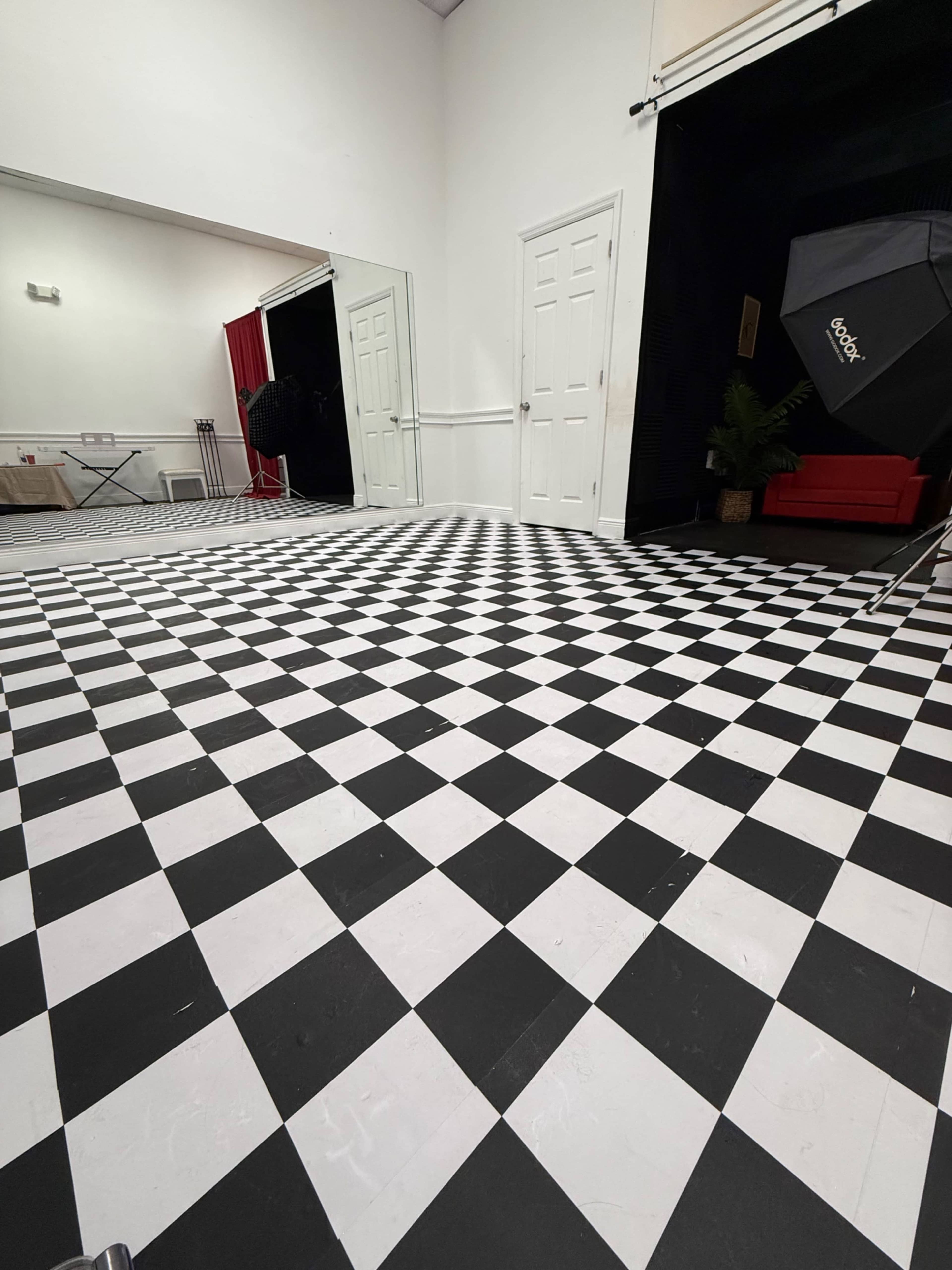 The image shows a spacious studio with a checkered black and white floor, mirrored walls, and two doors leading to different areas.