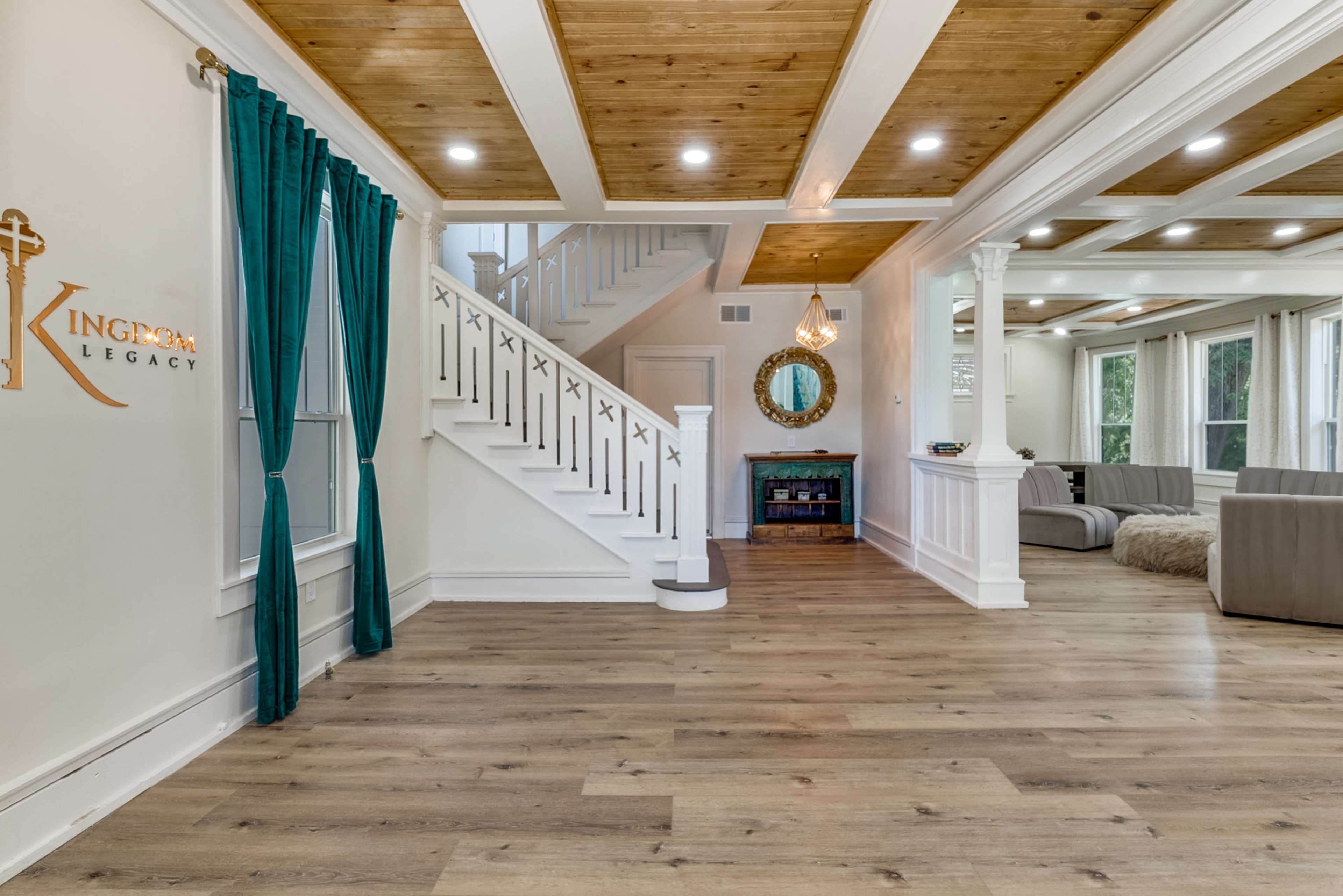 The image shows a modern interior with a staircase, a round mirror, and a living area featuring a sectional sofa and wooden ceiling beams.