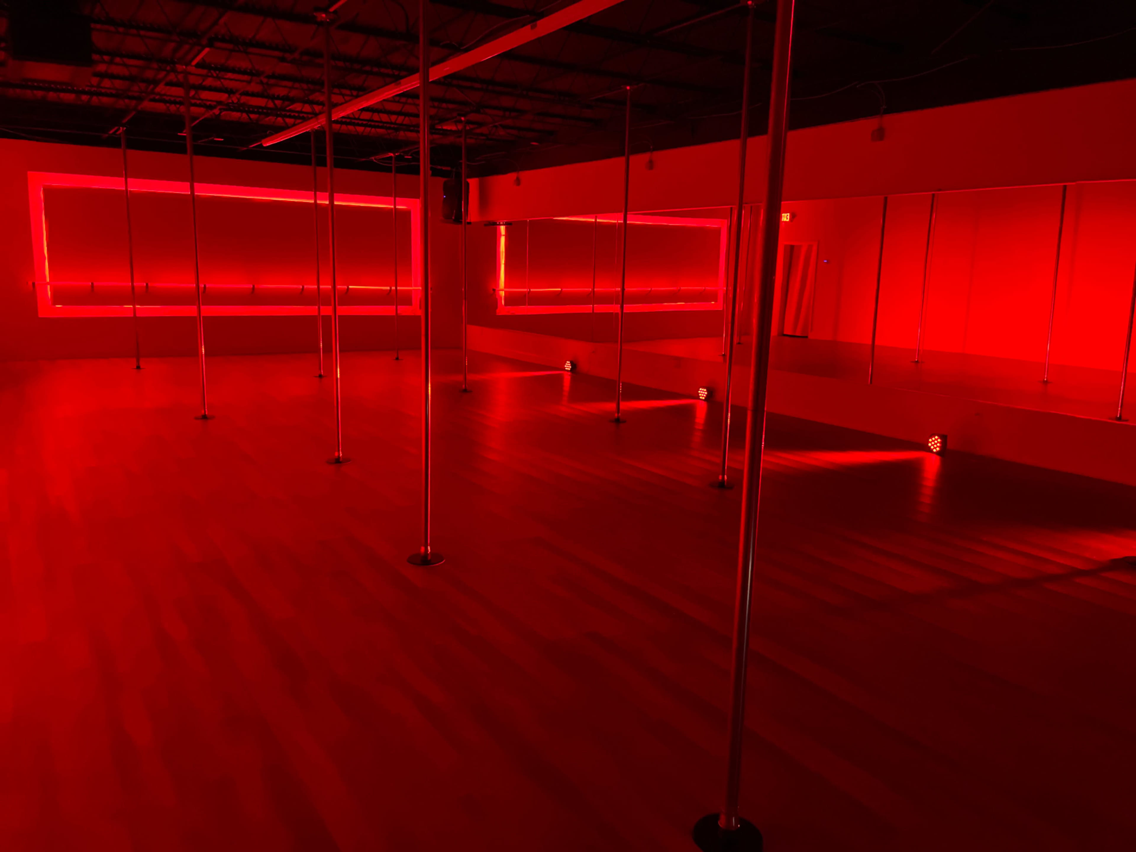 The image shows a dance studio with several pole dancing apparatuses illuminated by red lights.