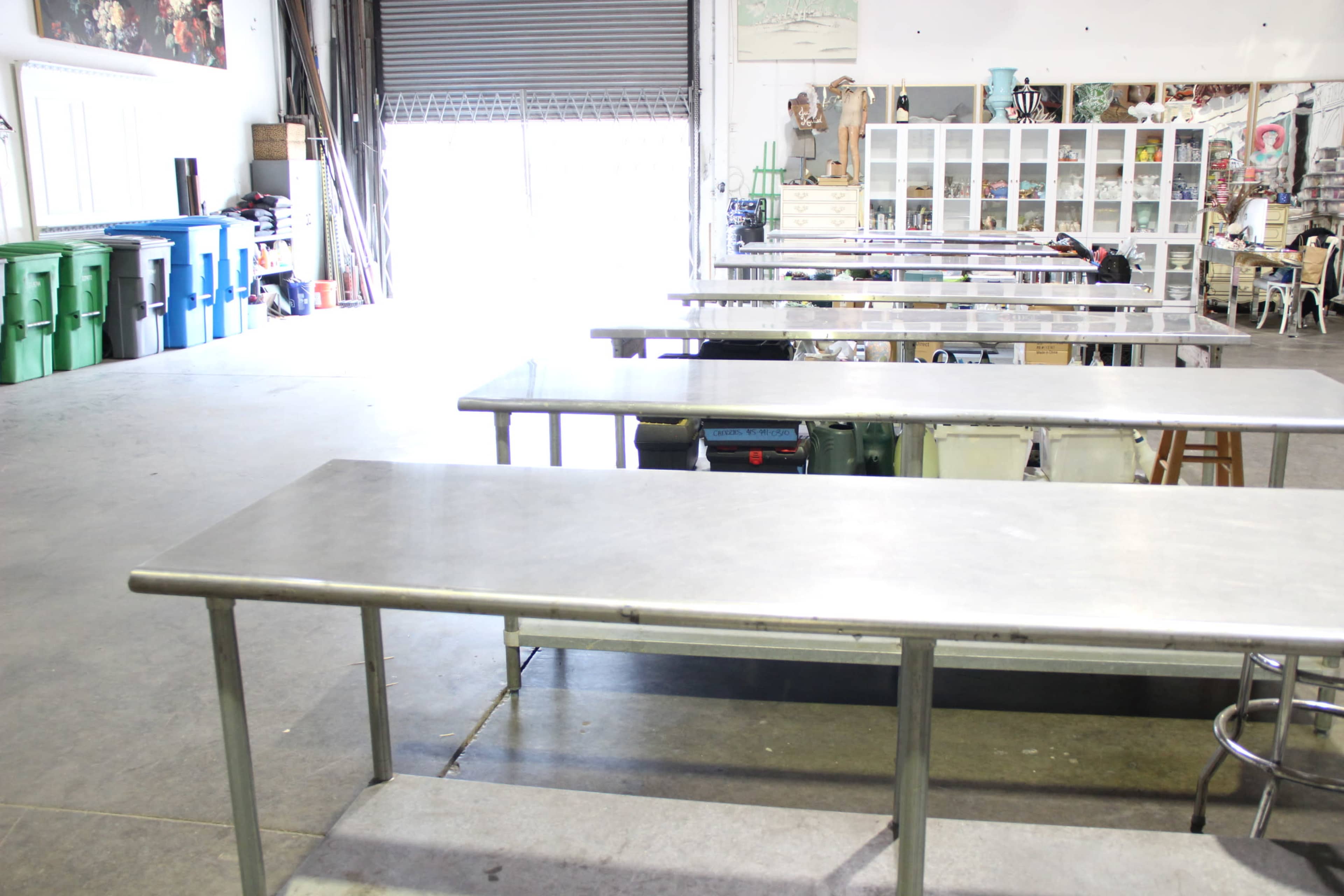 The image shows multiple stainless steel tables arranged in a spacious, industrial-style room with various storage bins and shelves in the background.