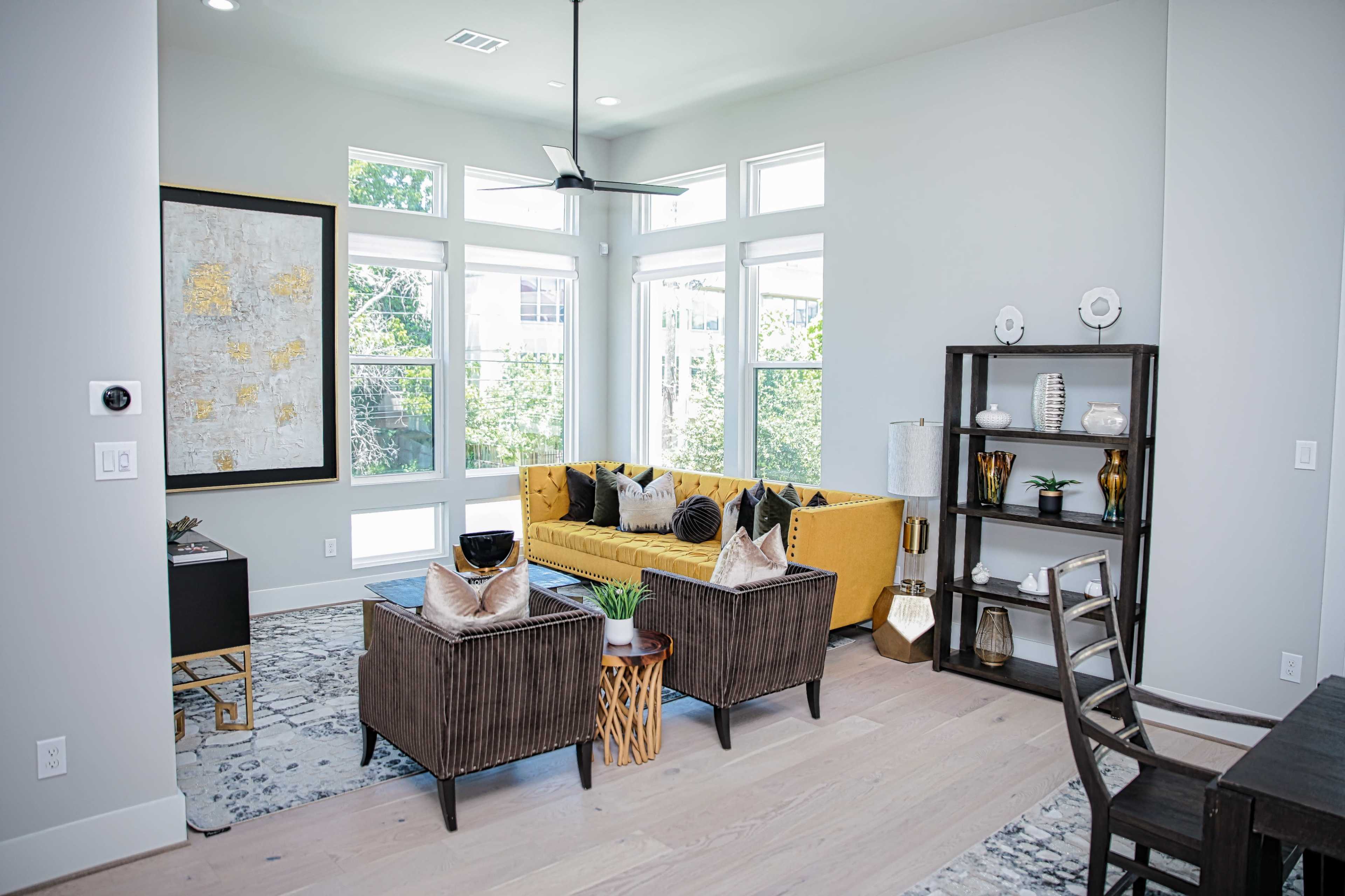 The living room features a yellow sofa, two dark brown armchairs, and a wooden shelf, with large windows allowing natural light to fill the space.