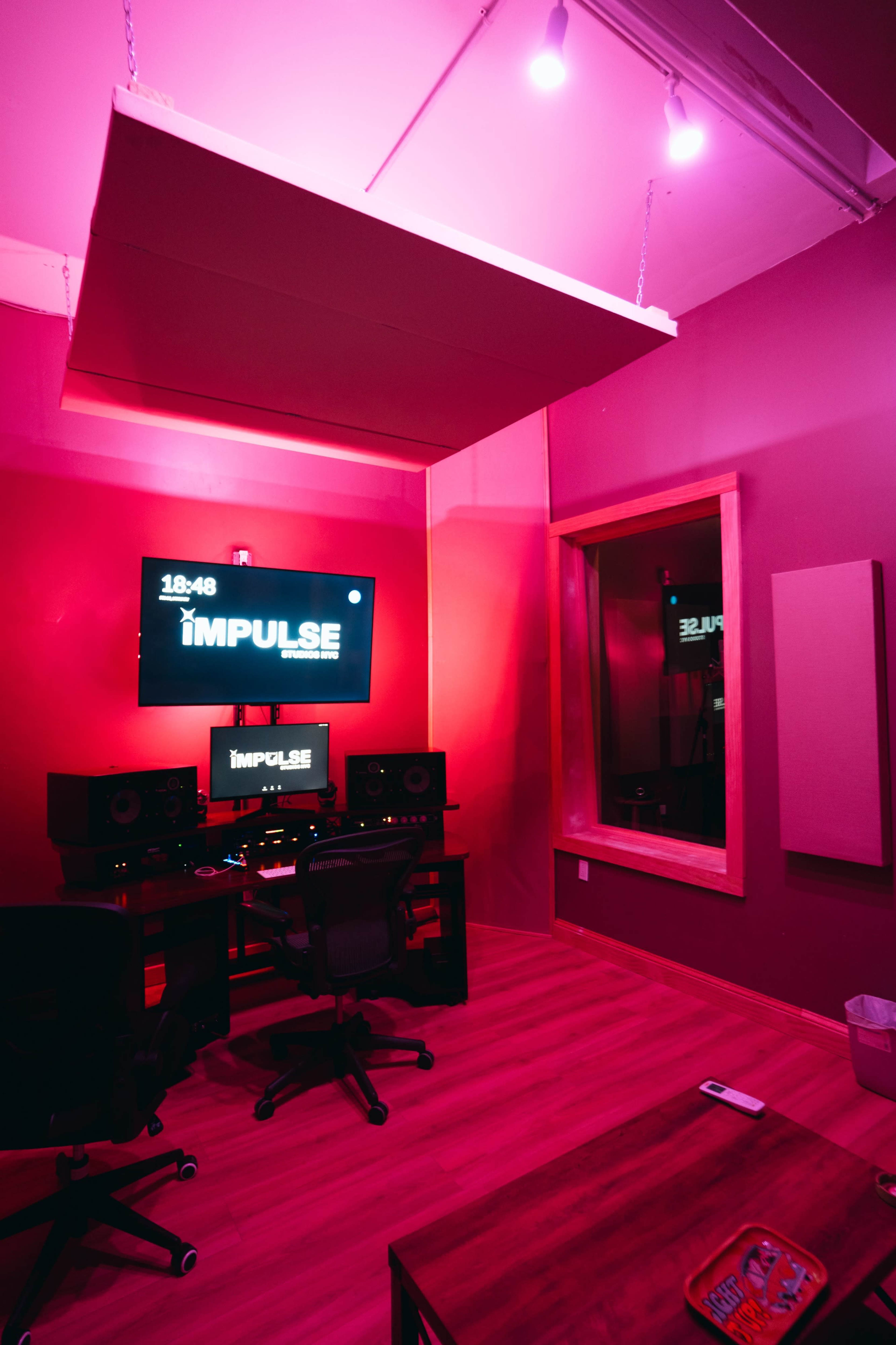 A modern music studio features a dark pink interior, a large monitor displaying "IMPULSE," and audio equipment arranged around a central workstation.