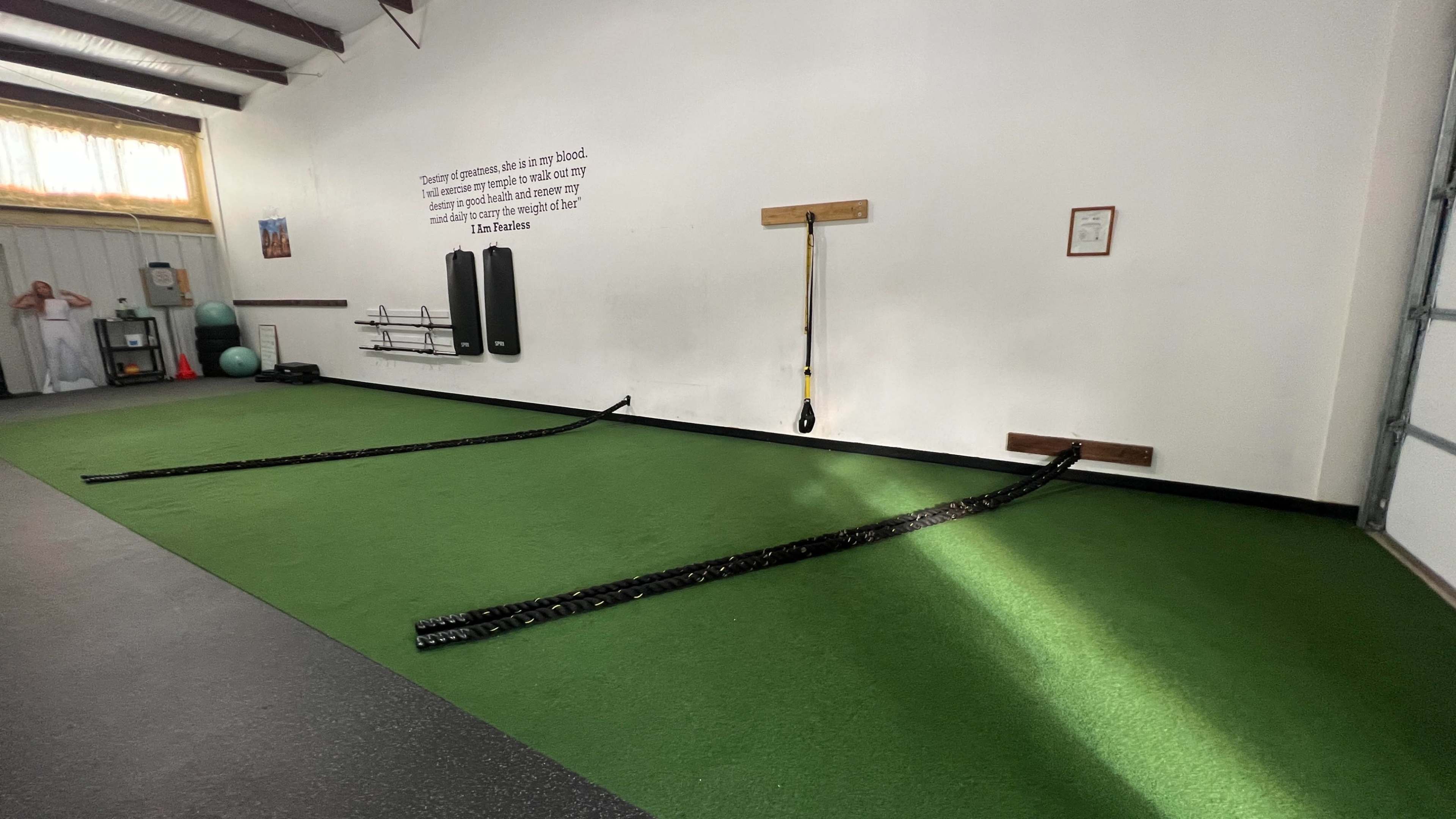 A fitness area with green artificial turf, featuring two black rubber training sled tracks along the floor and various exercise equipment mounted on the wall.