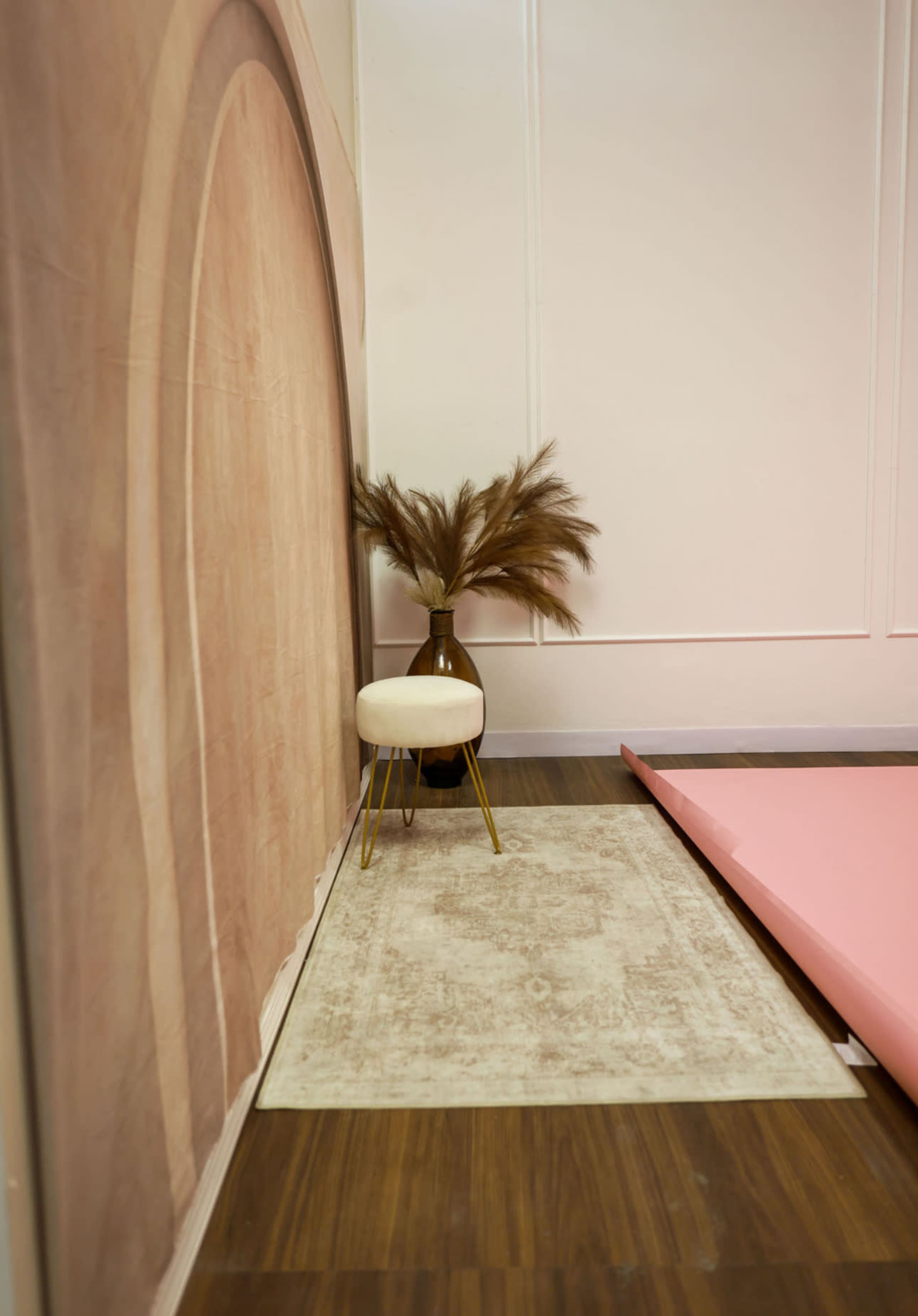 The scene features a small, modern space with a beige backdrop, a white upholstered chair with gold legs, a vase holding dried plants, and a light pink platform on the floor.