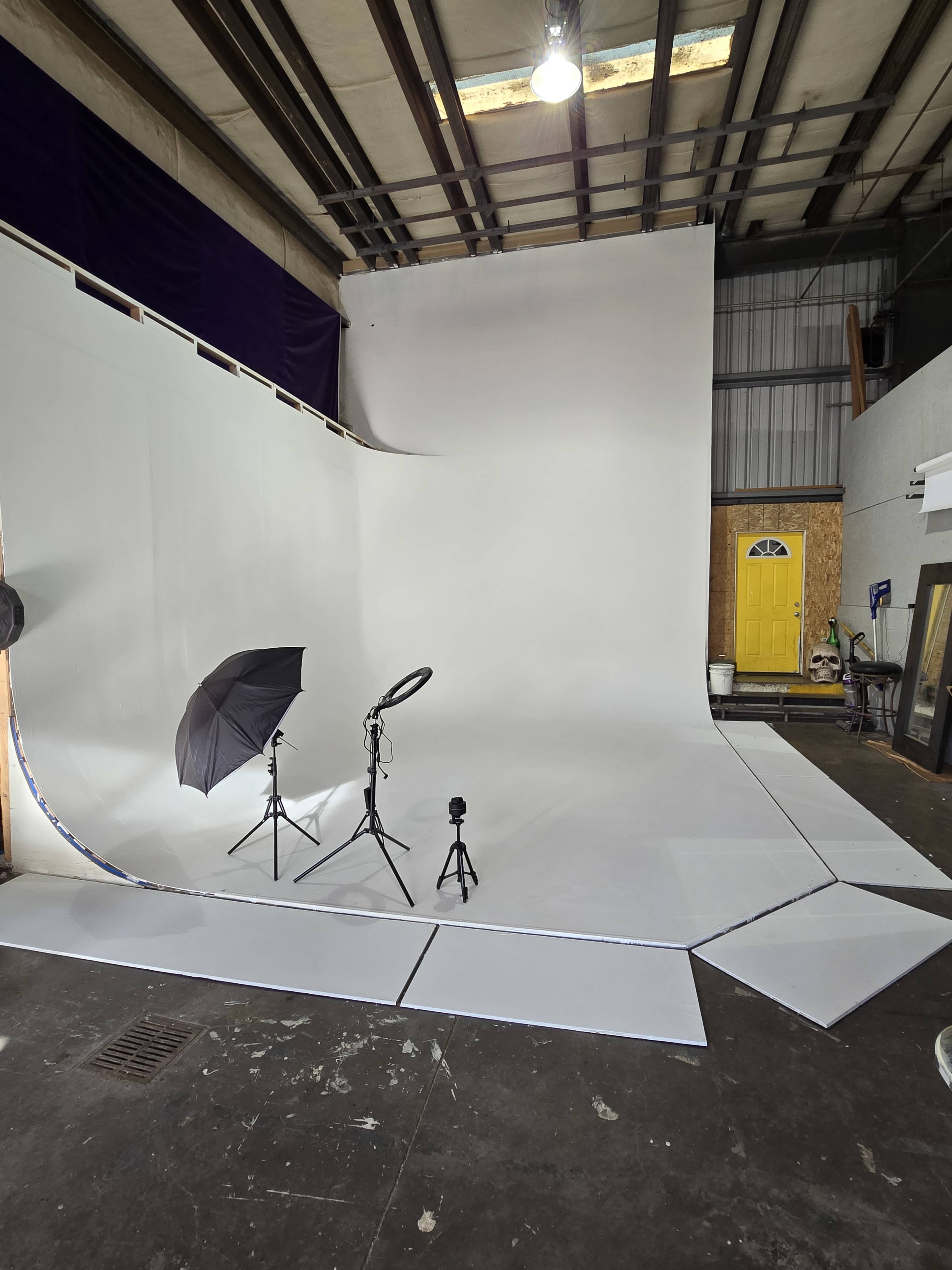 The image shows a photography studio with a seamless white backdrop, two light stands, and a yellow door in the background.