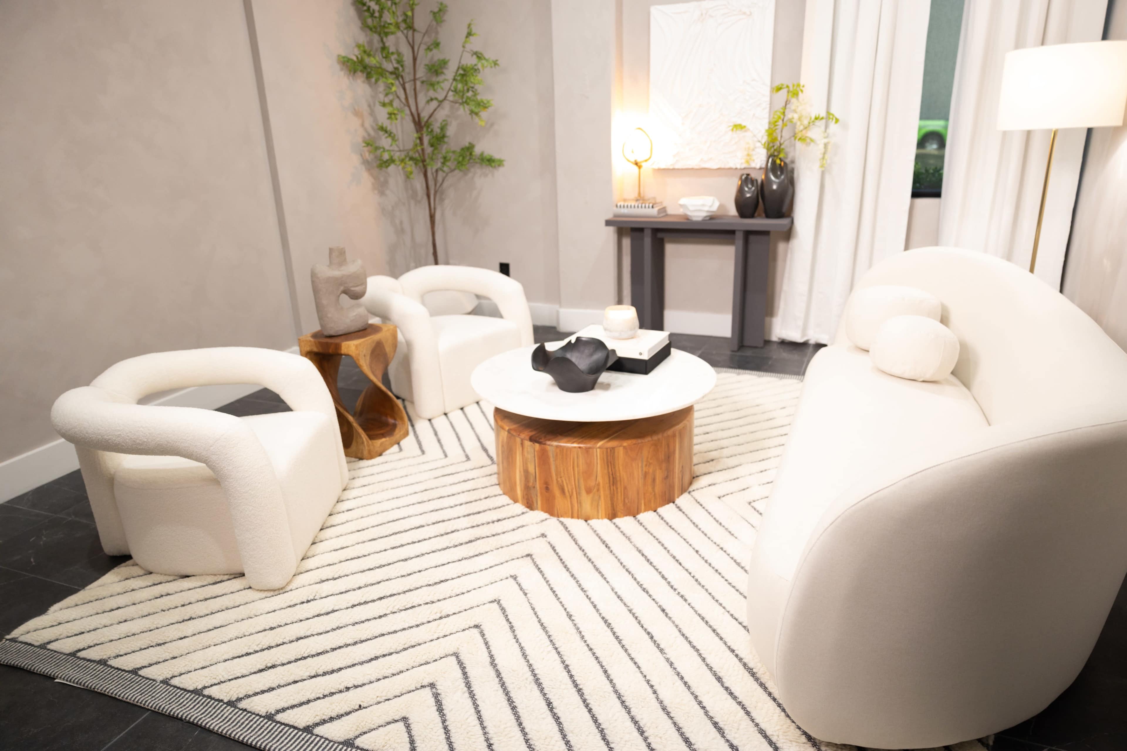 A cozy seating area features two white upholstered chairs and a round wooden coffee table on a patterned rug, surrounded by minimal decor and natural light.