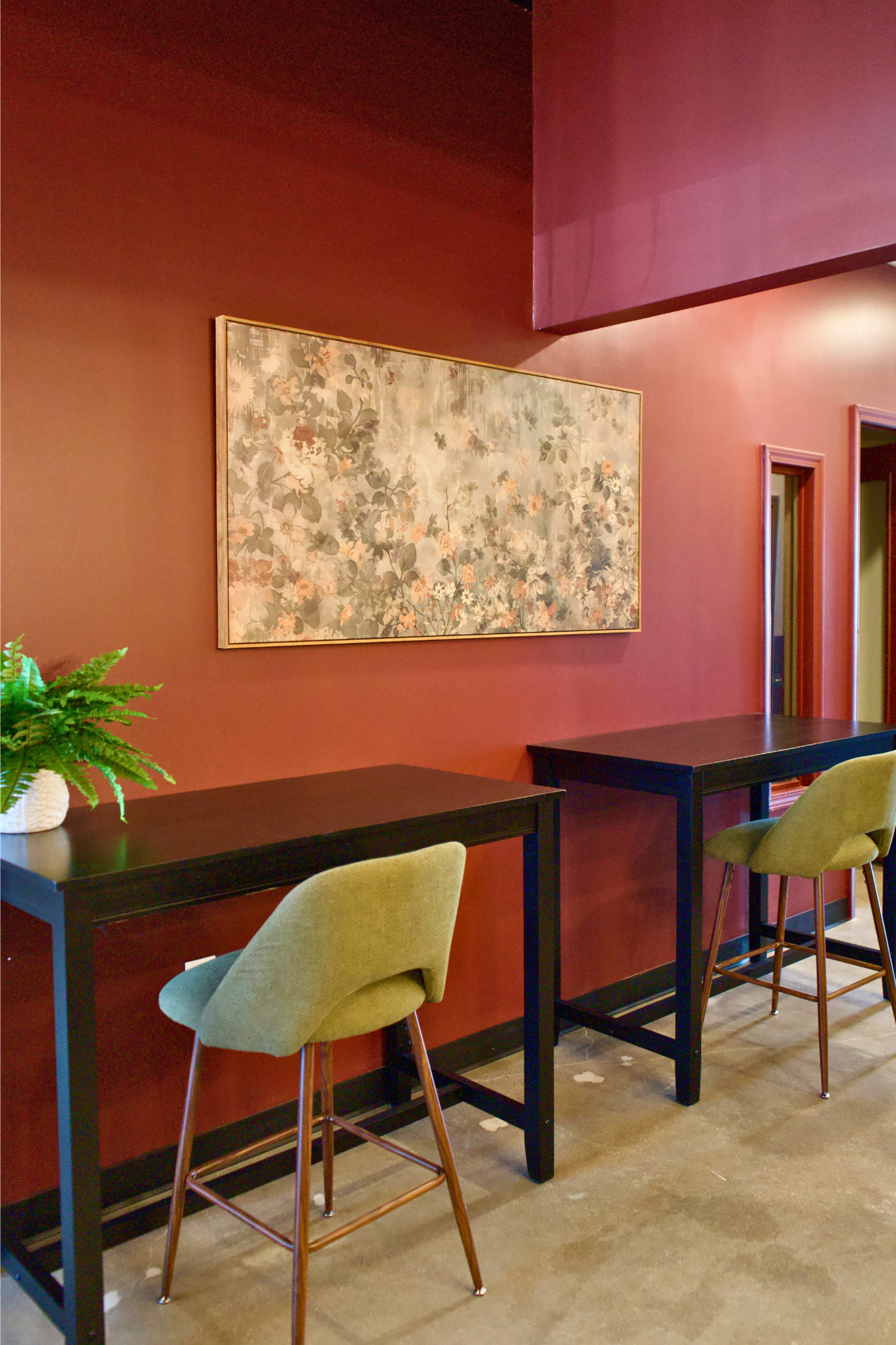The image shows a modern interior featuring two high tables with green upholstered stools against a dark red wall, adorned with a large floral artwork.