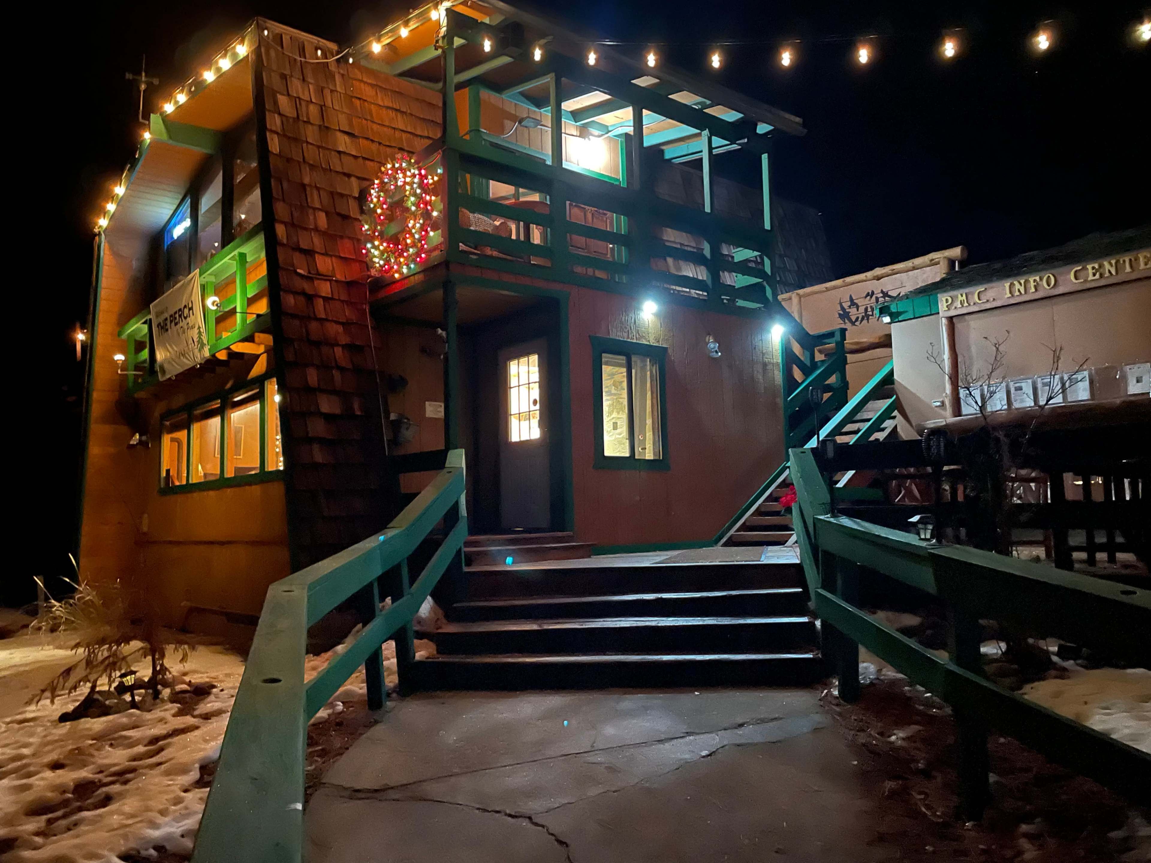 A two-story building with green railings and Christmas lights is illuminated at night, surrounded by patches of snow.