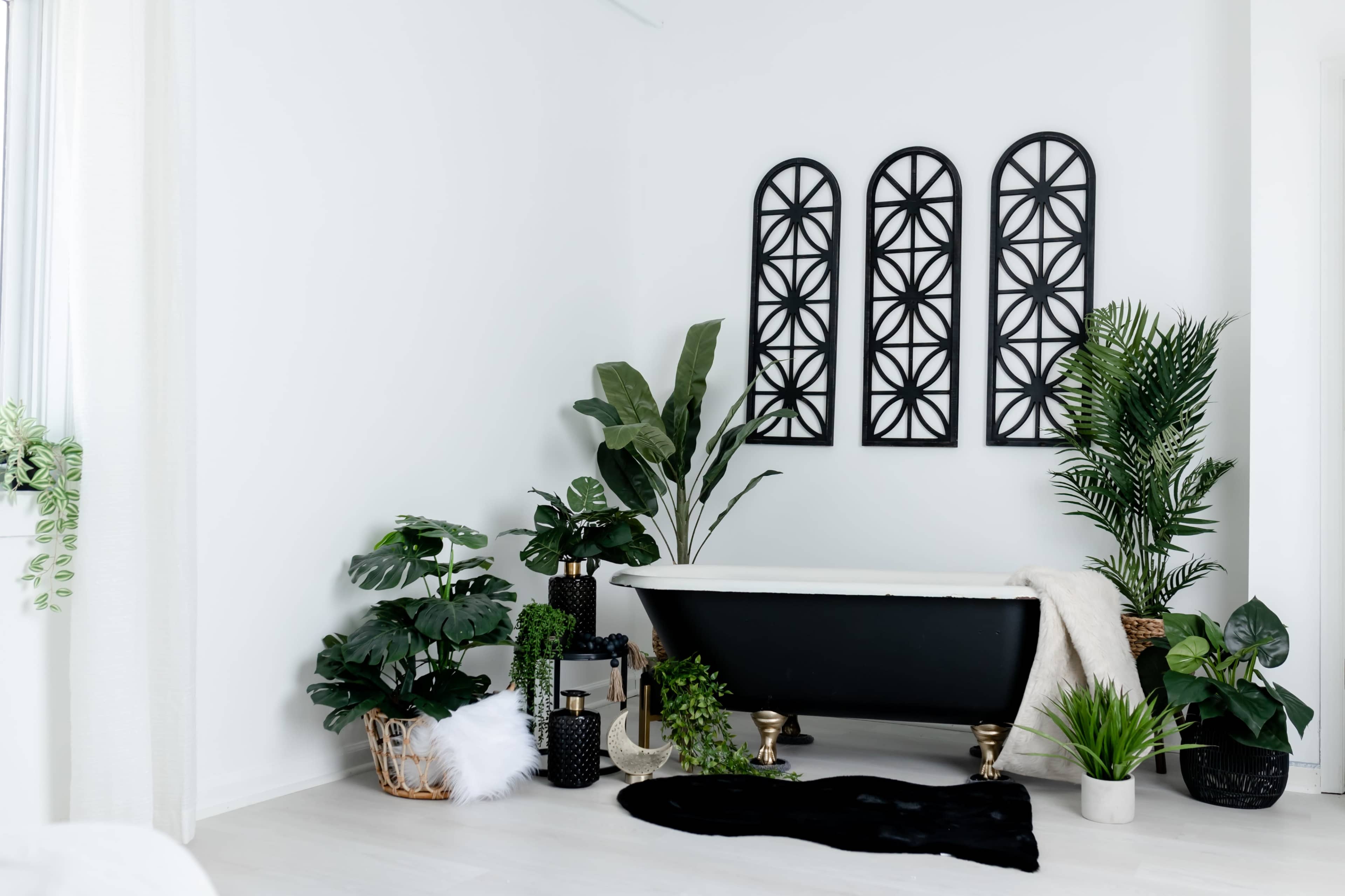 A black freestanding bathtub is surrounded by various plants and decorative elements against a white wall with three arched mirrors.