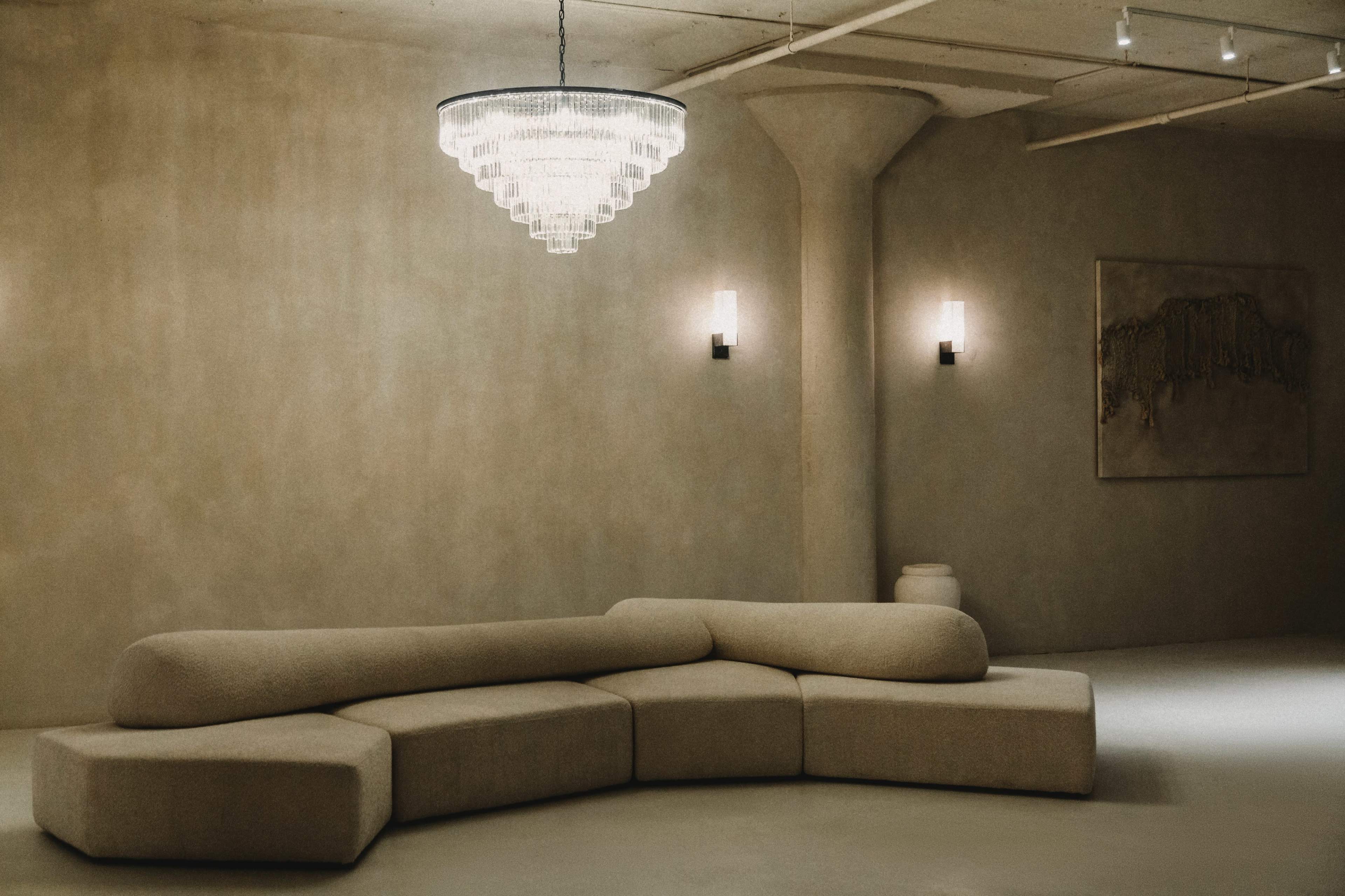 A minimalist interior with a curved beige sofa, a large chandelier hanging from the ceiling, and simple wall sconces providing light.